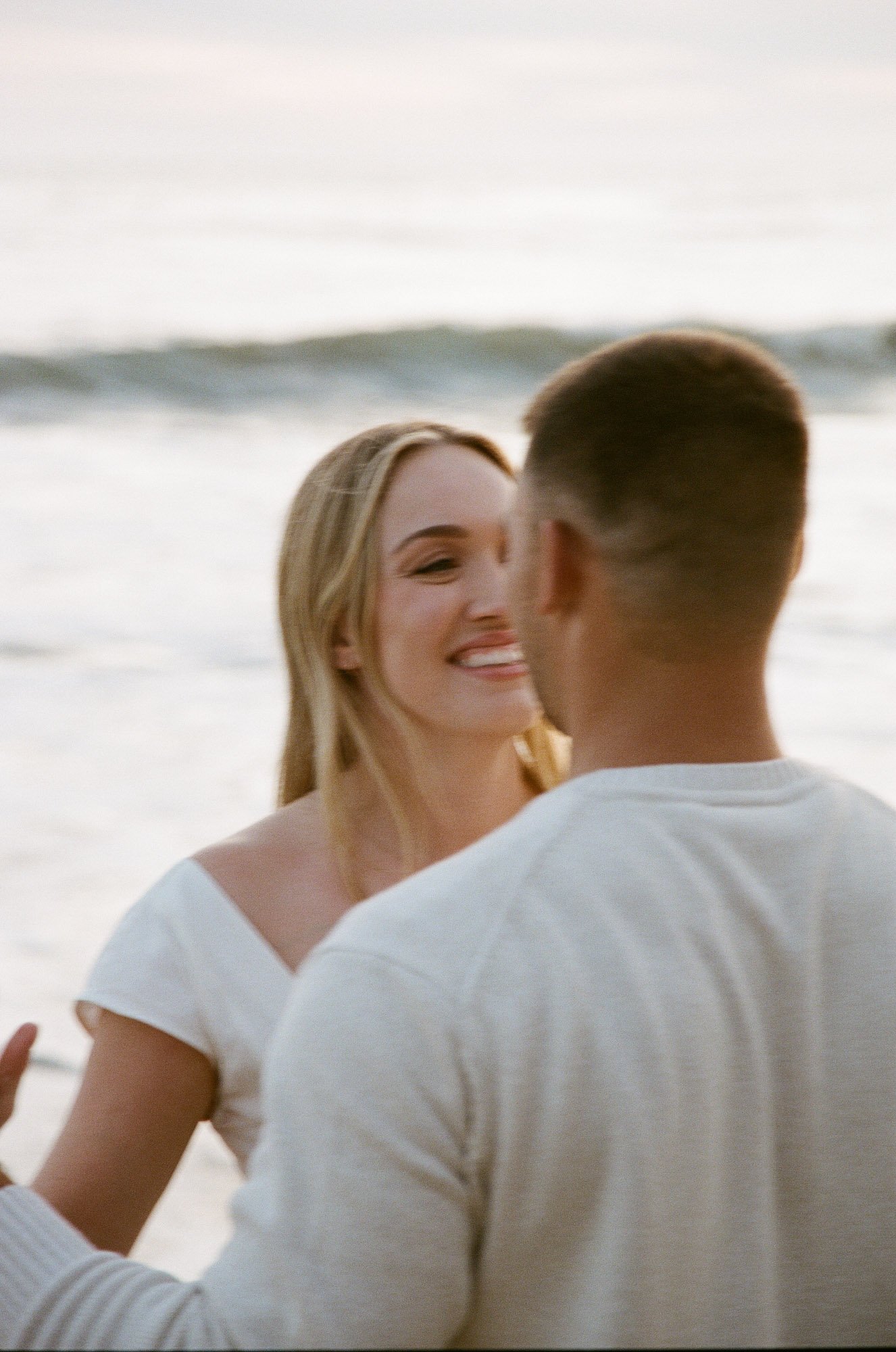 A smiling woman with blonde hair and a man with short brown hair face each other on a beach with ocean waves in the background.