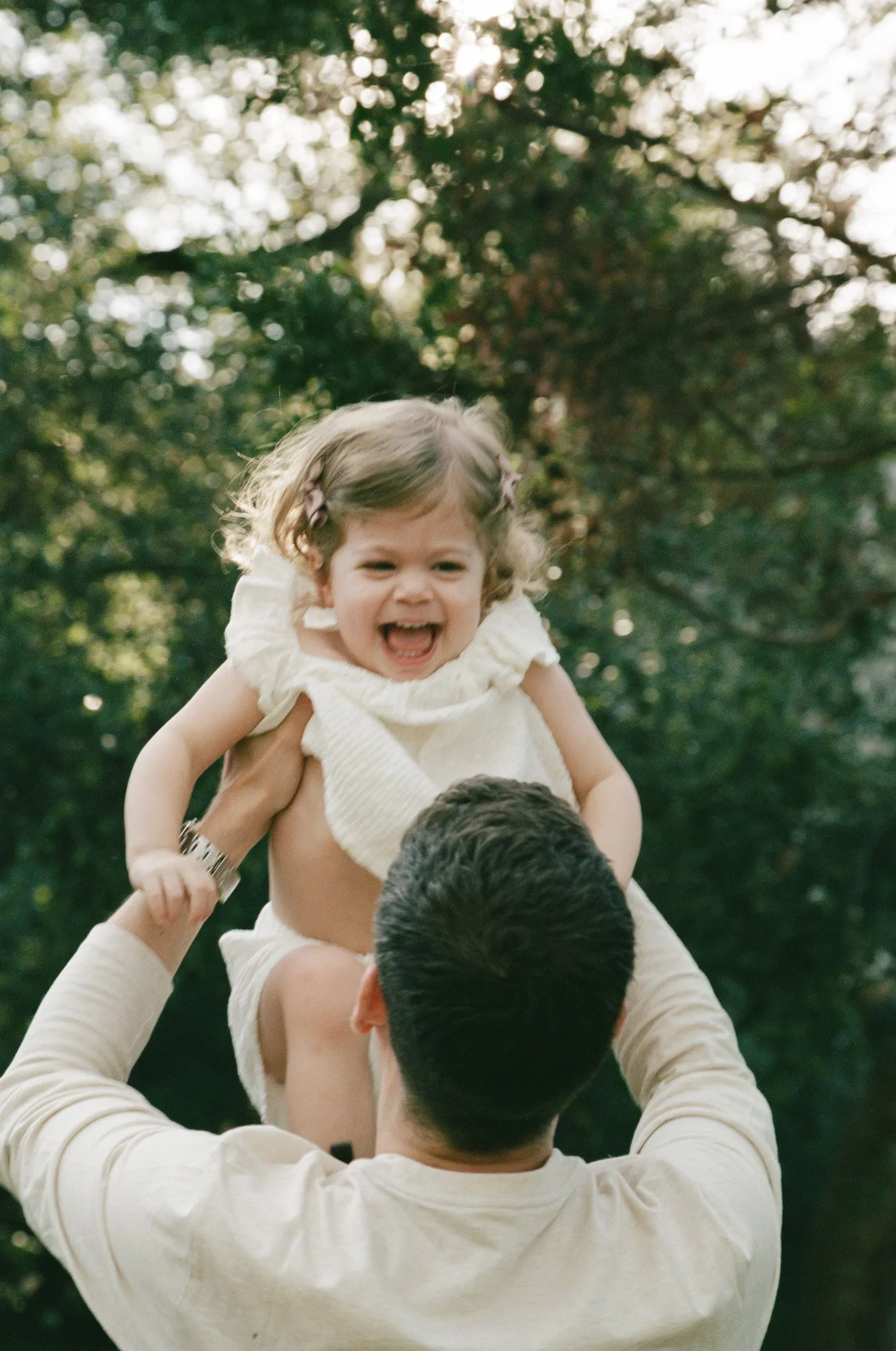 A man lifting a young girl on his shoulders outdoors with trees in the background. The girl is laughing and smiling.