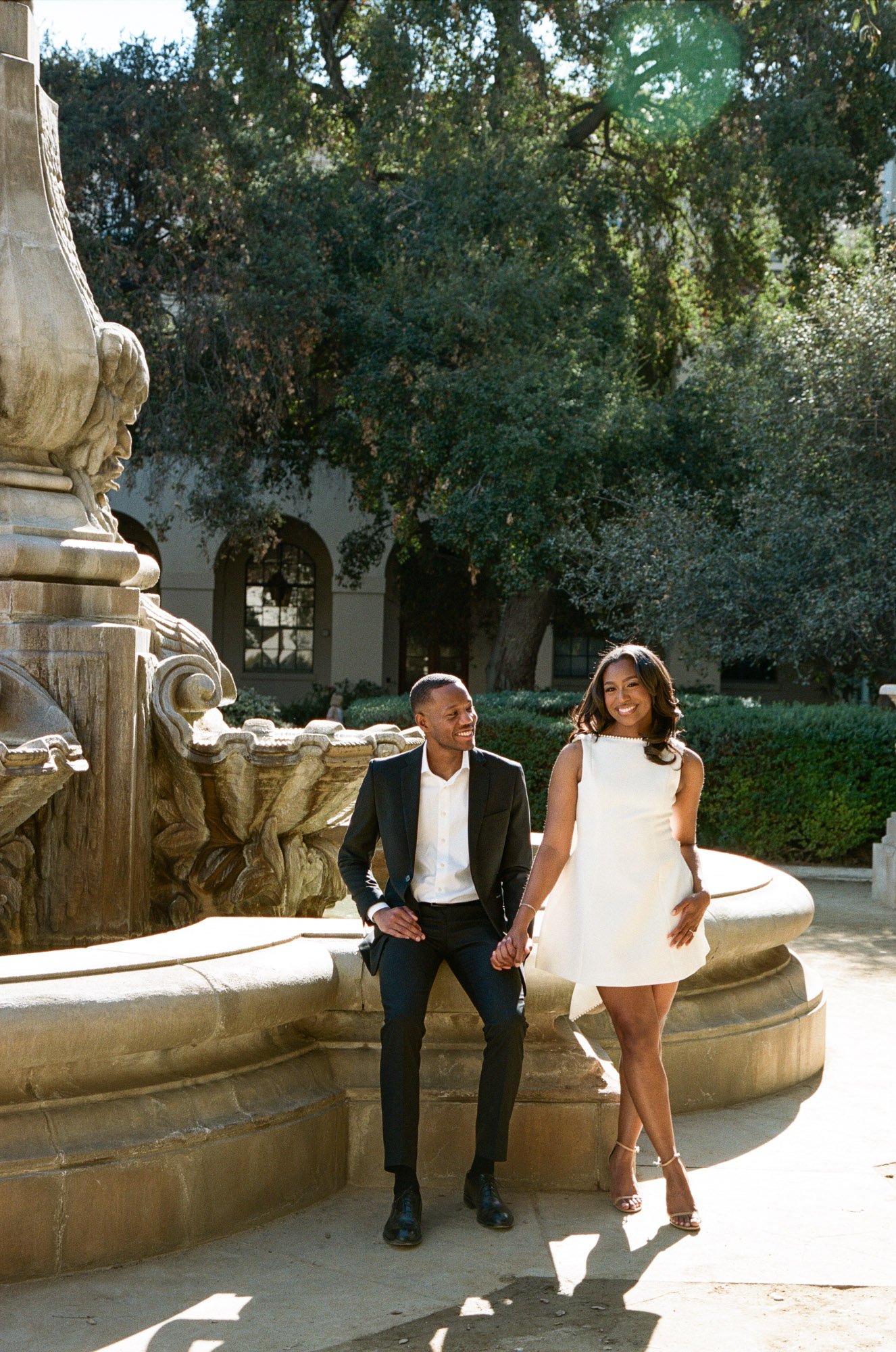 A smiling man in a black suit and dress shoes, and a smiling woman in a white dress and heels, standing together holding hands in front of a large ornate fountain with trees and a building in the background.