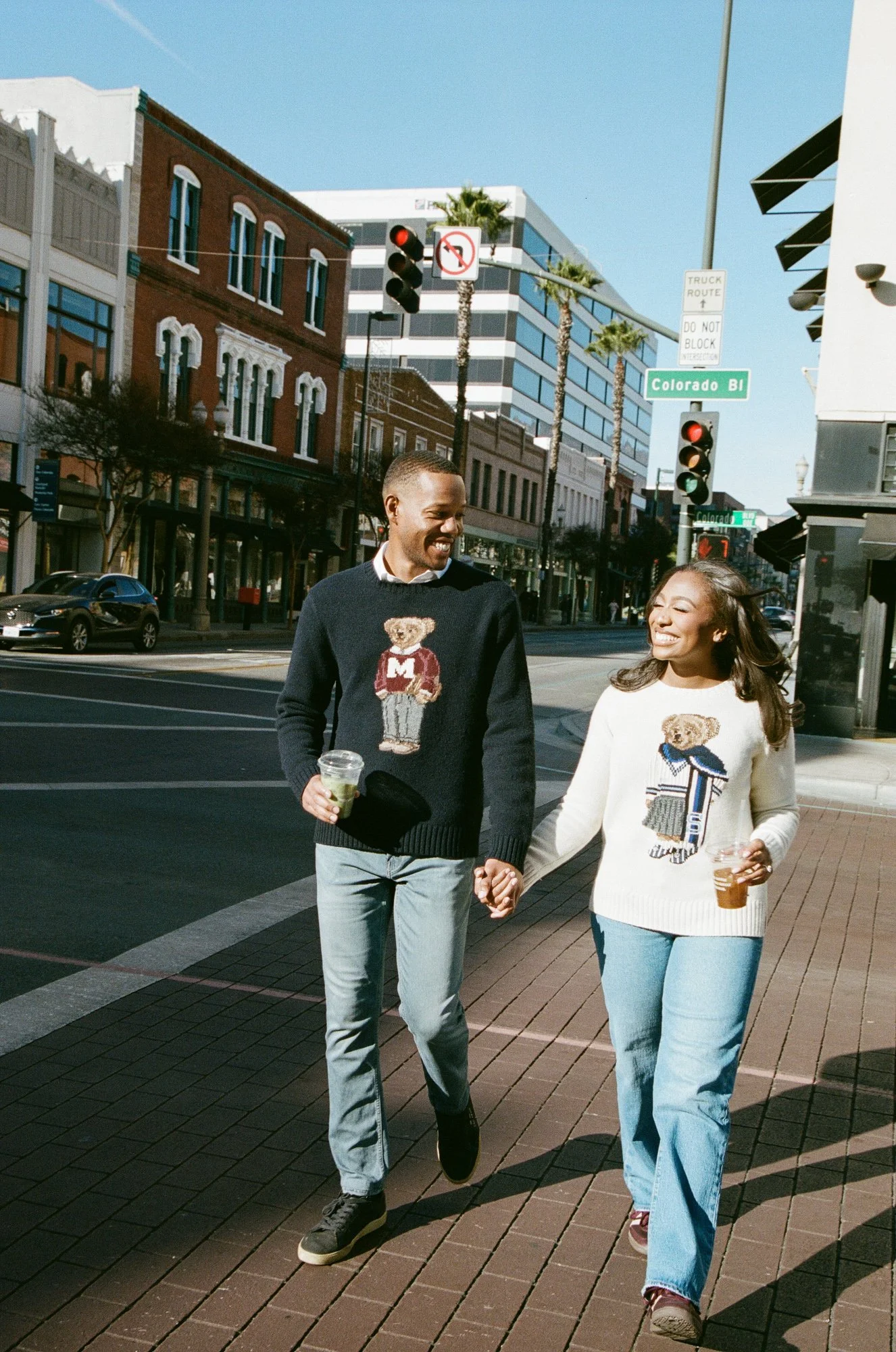 A smiling man and woman walking hand-in-hand on a city sidewalk, each holding a drink, with buildings and palm trees in the background, sunny day.