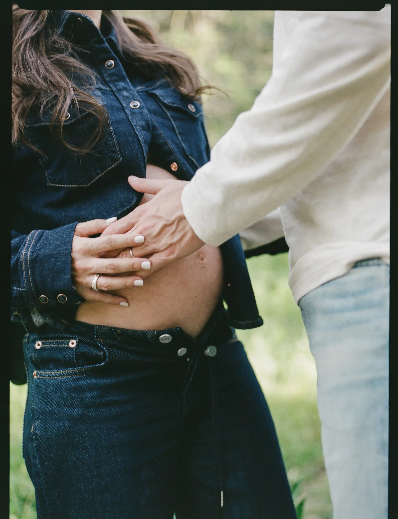 A pregnant woman wearing a denim jacket and black jeans is holding her belly, while a man in a beige shirt and jeans is gently touching her belly in an outdoor setting.