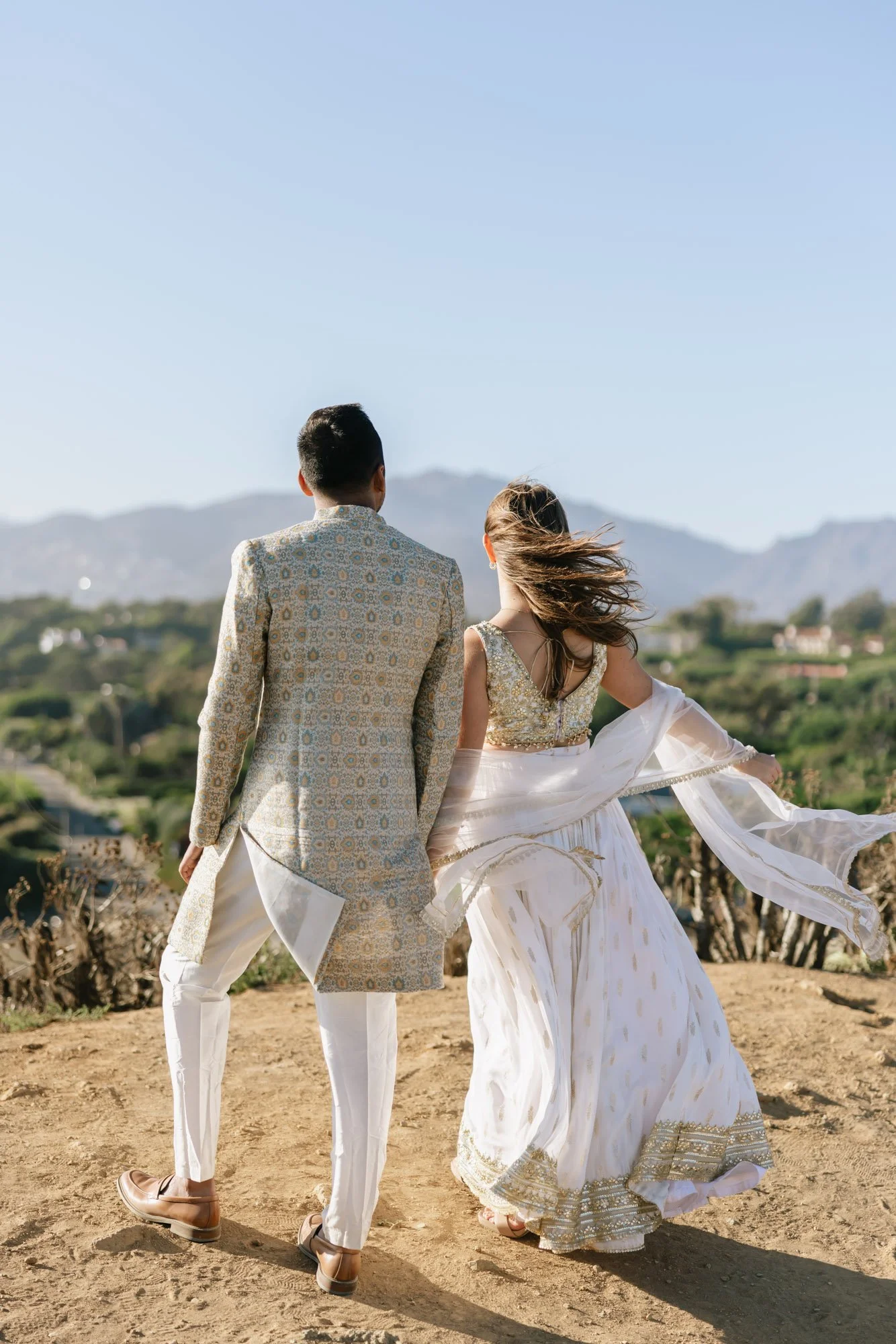 A couple dressed in traditional Indian wedding attire standing outdoors on a dirt path with mountains in the background. The woman is wearing a cream and gold lehenga with flowing fabric, and the man is in a beige patterned sherwani with white trouse