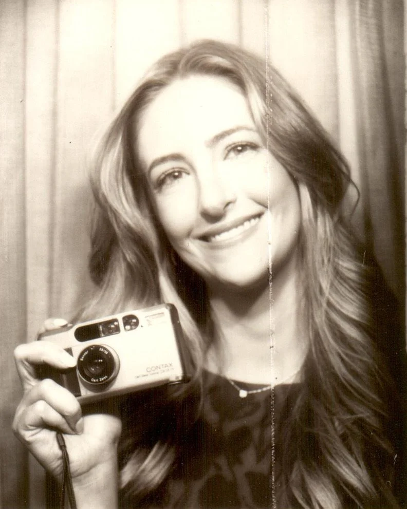 Black and white photo of a smiling woman with long wavy hair holding a vintage camera.