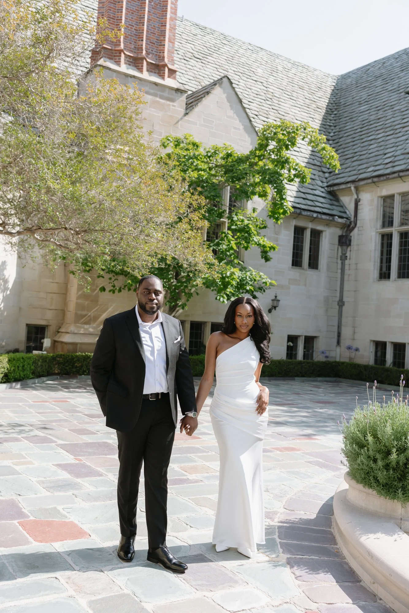A couple holding hands and walking outdoors in front of a large stone building with a steep roof and tall brick chimney. The man is wearing a black suit with white shirt, and the woman is wearing a white strapless wedding gown. They are walking on a 