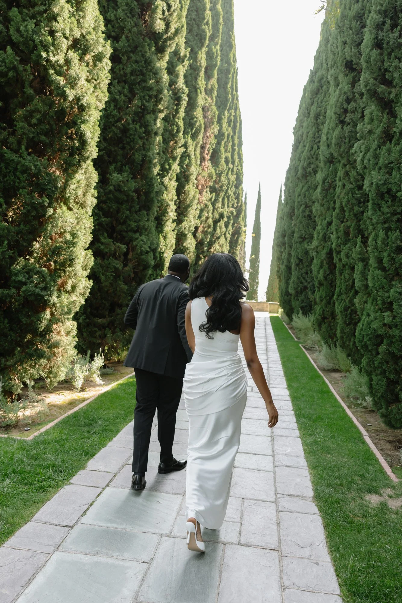 A woman in a white wedding dress walking on a stone pathway through a garden with tall, neatly trimmed green trees on each side, with a man in a black suit walking ahead of her.