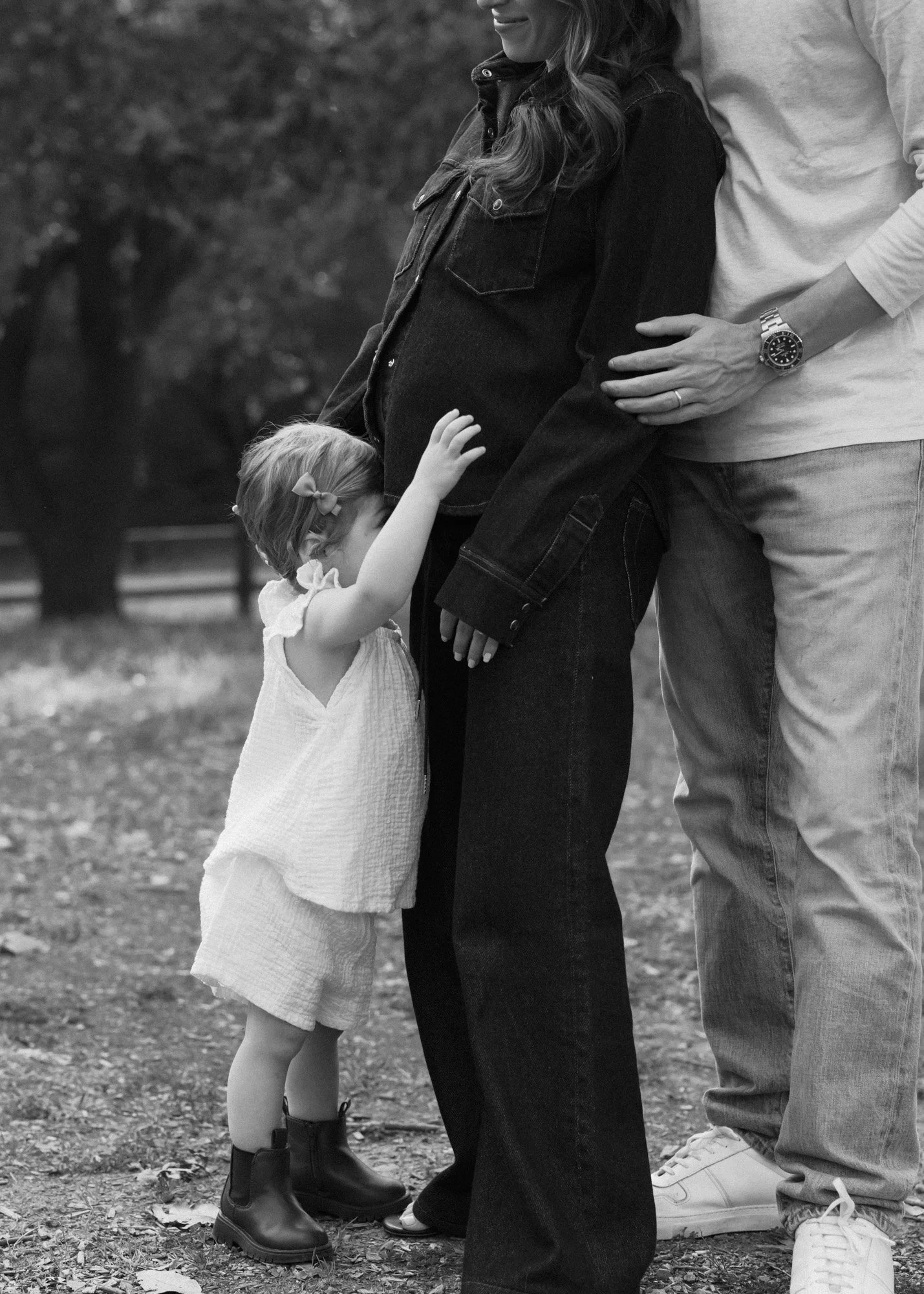 A family of three standing outdoors. A young girl with a bow in her hair is gently touching a pregnant woman's belly, and a man is standing beside her with his hand on her back. The pregnant woman is smiling slightly, and their heads are close togeth