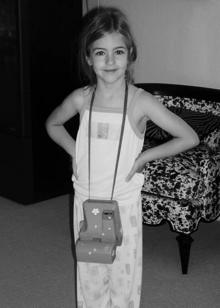 A young girl standing indoors with hands on hips, wearing pajamas and a necklace with a large decorative pendant, smiling at the camera.