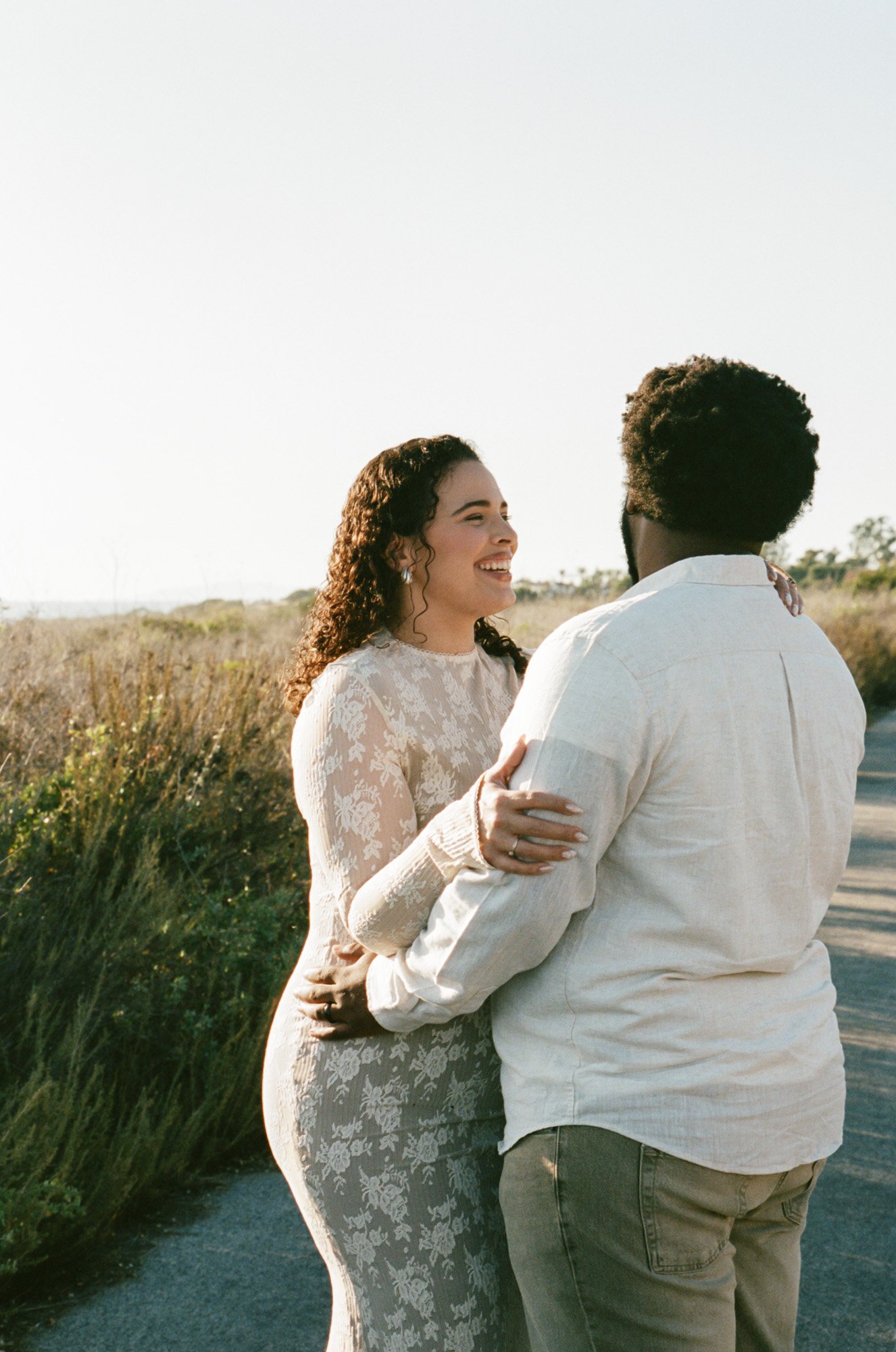 A woman and a man embracing outdoors on a sunny day, smiling and looking at each other with a background of tall grass and trees.