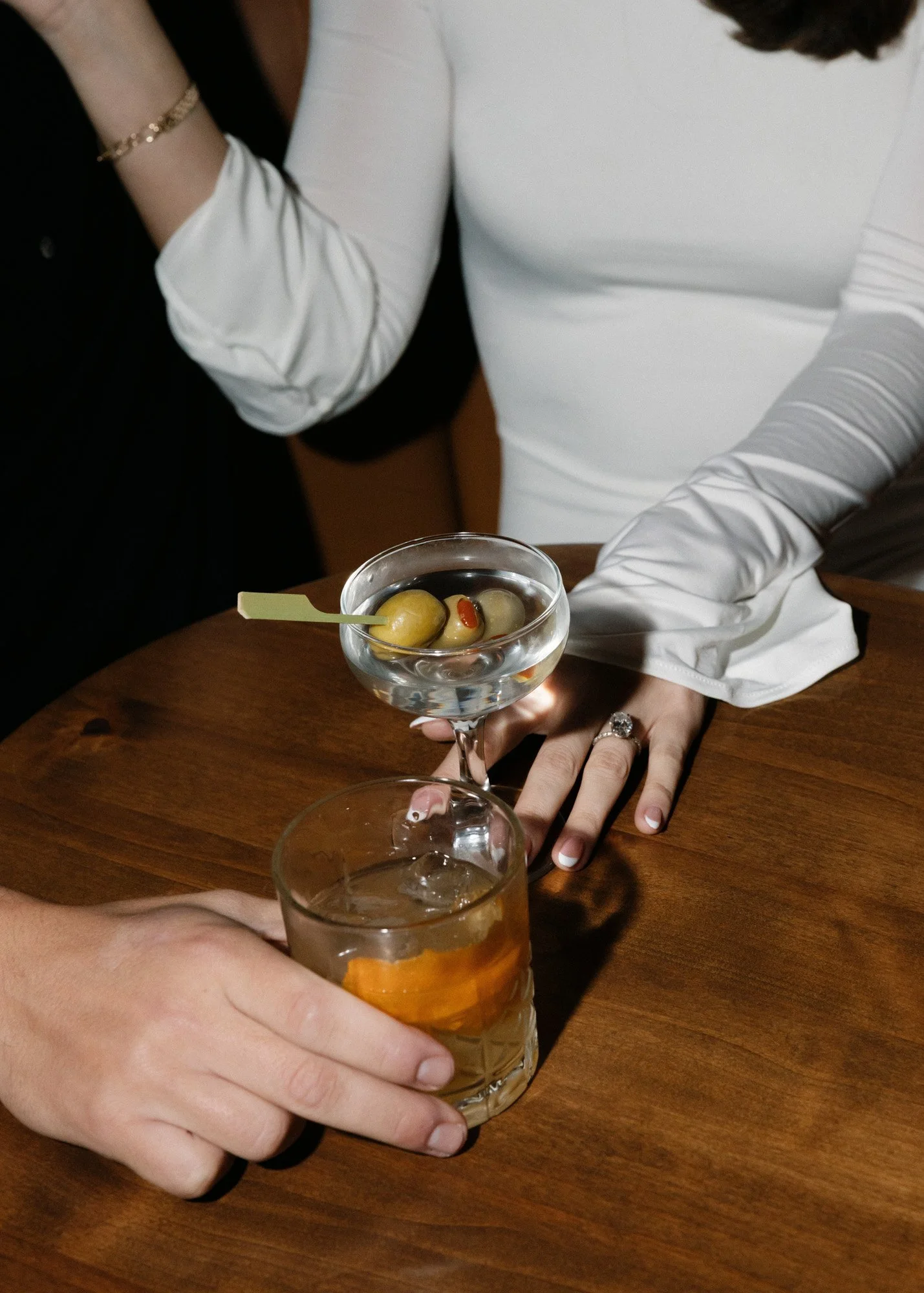 Two women at a table, one holding a glass of drink with ice, the other holding a cocktail glass with olives. They are dressed in stylish white and black outfits, and their hands are resting on a wooden table.