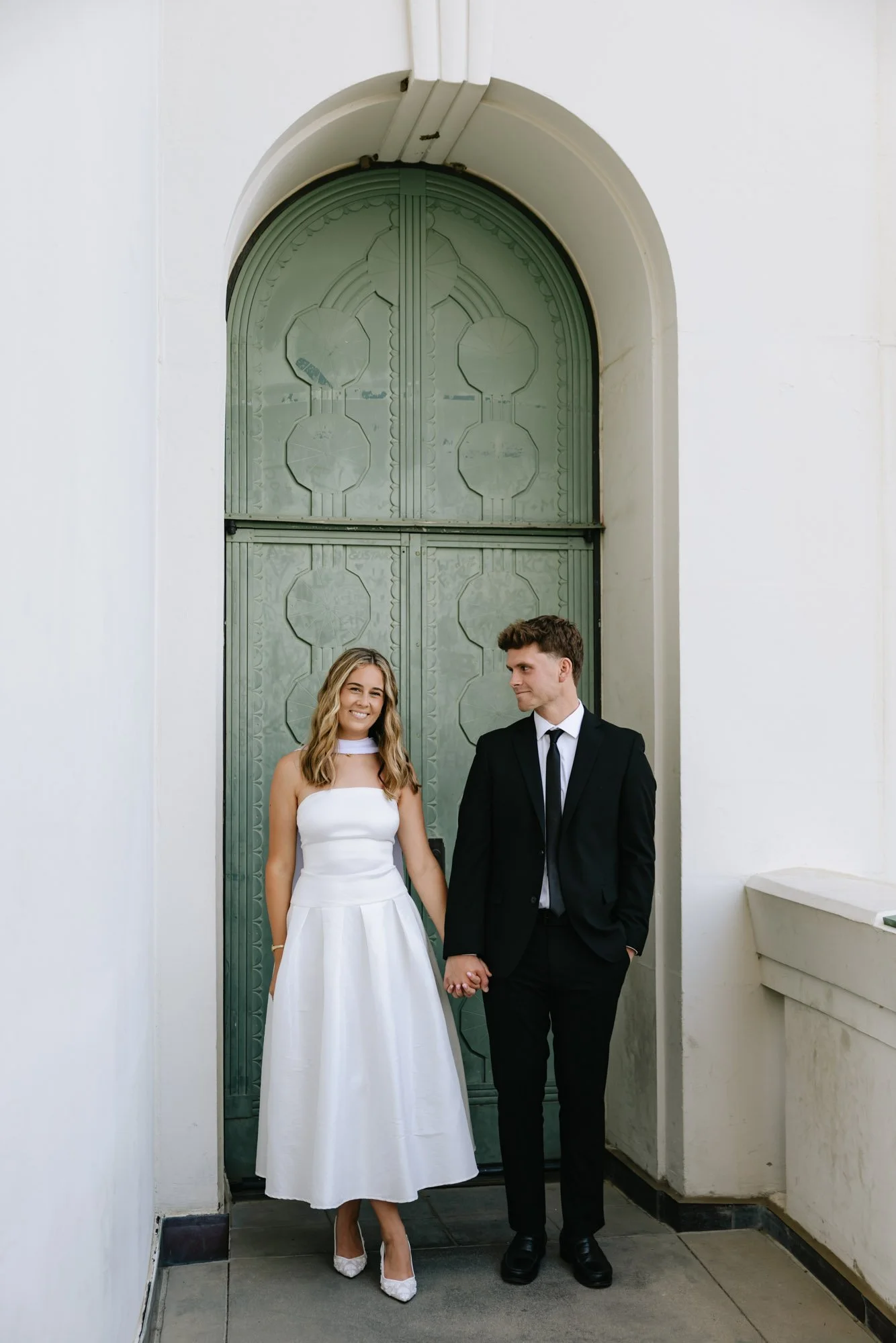 A bride and groom holding hands, standing in front of a large green arched door. The bride is wearing a white strapless dress and heels, with long blonde hair. The groom is dressed in a black suit with a white shirt and black tie, with short brown ha