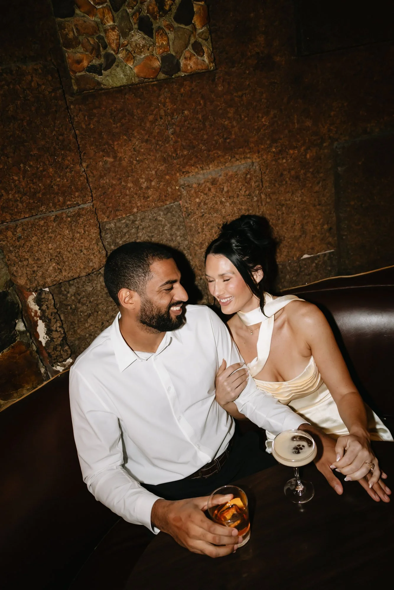 A couple sitting closely together in a dimly lit bar or restaurant, smiling and enjoying drinks, with a textured stone wall in the background.