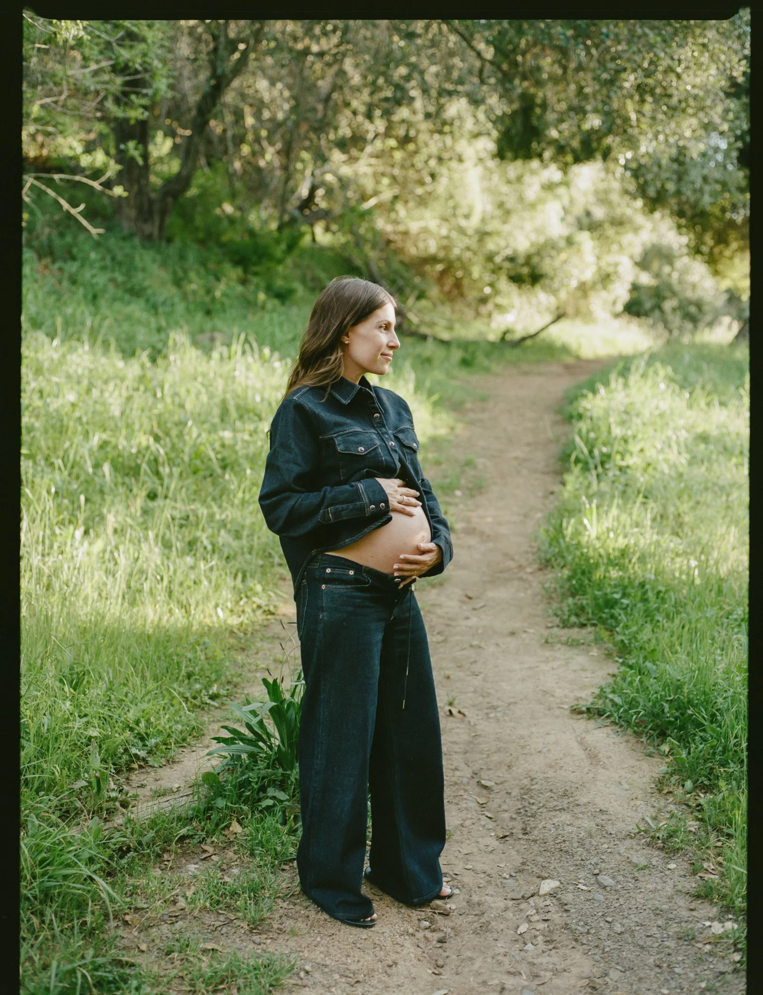 A woman with long brown hair, wearing a black denim jacket and wide-leg jeans, stands on a dirt path in a lush green forest, gently holding her pregnant belly with both hands and looking to the side with a peaceful expression.