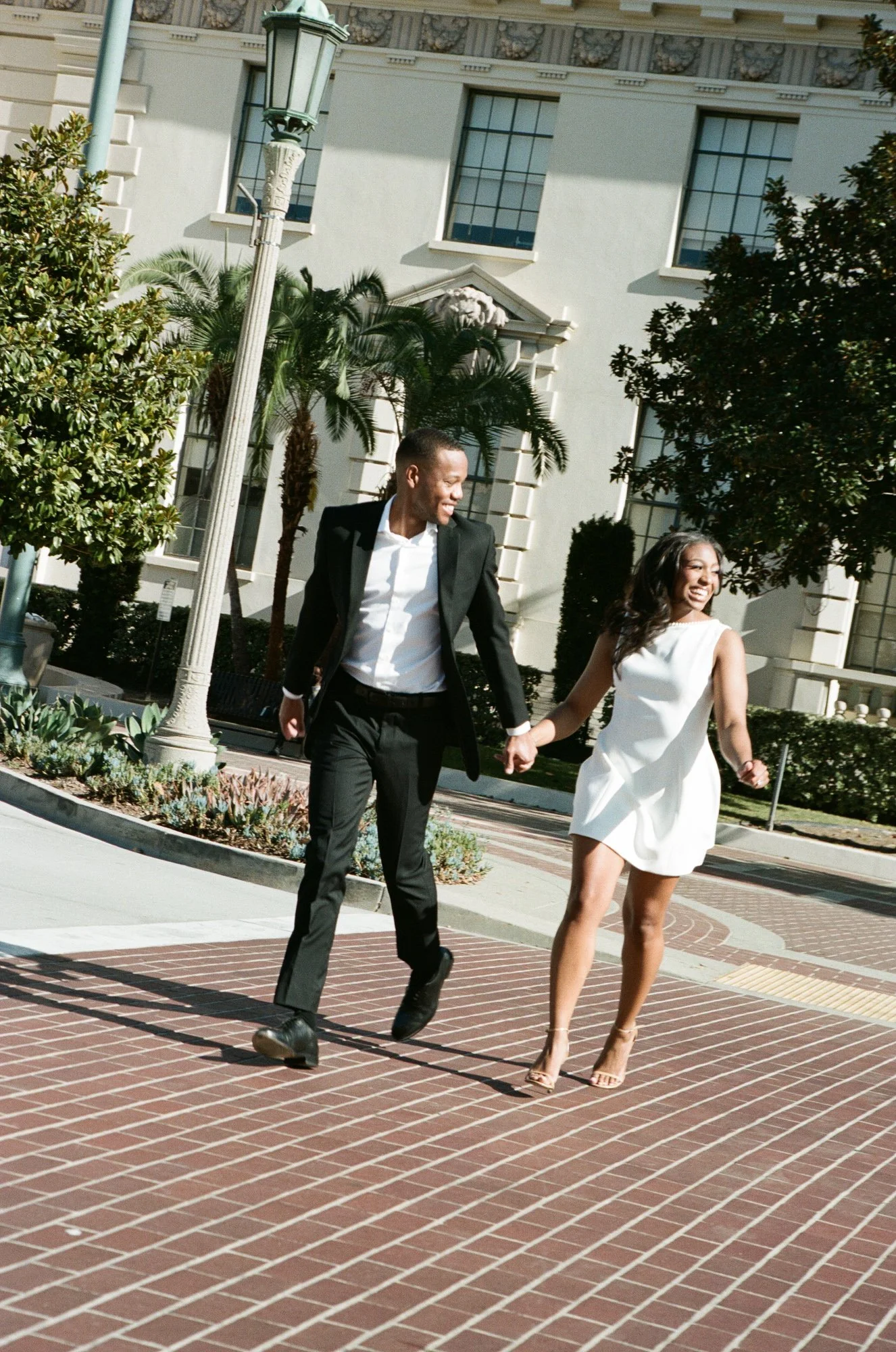 A smiling couple, a man in a black suit and a woman in a white dress, holding hands and walking on a brick sidewalk outdoors, with trees, bushes, and a white building with large windows in the background.