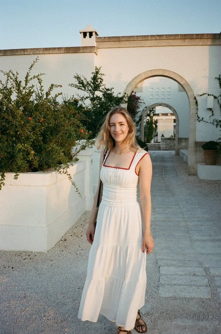 A young woman in a white summer dress standing outside near a white wall and archway, smiling, with greenery in the background during daytime.