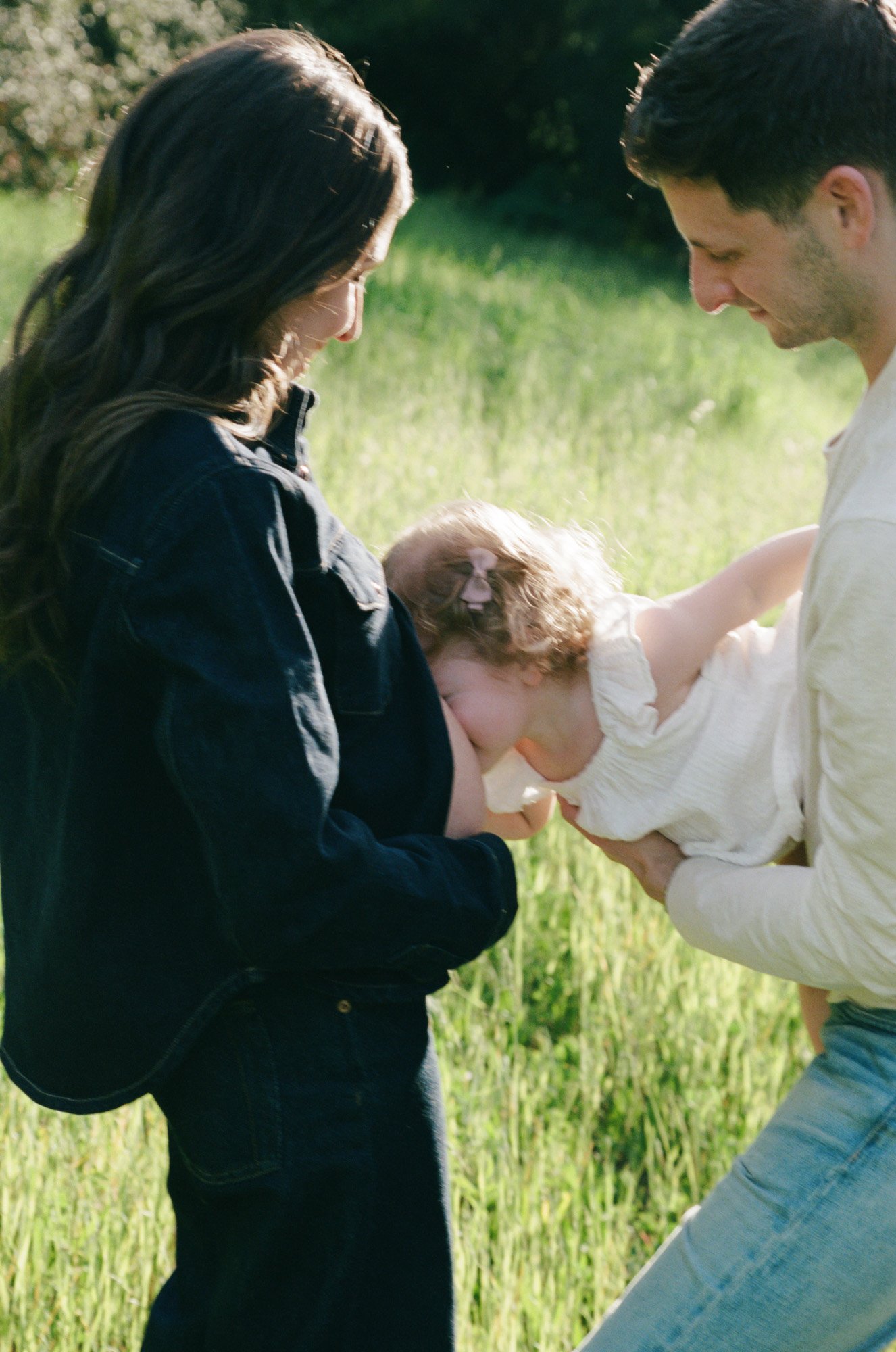 A man and a woman are outdoors in a grassy field, with a young girl between them. The girl is leaning into the man's chest as they all look downward, and they are smiling gently, with their foreheads touching.