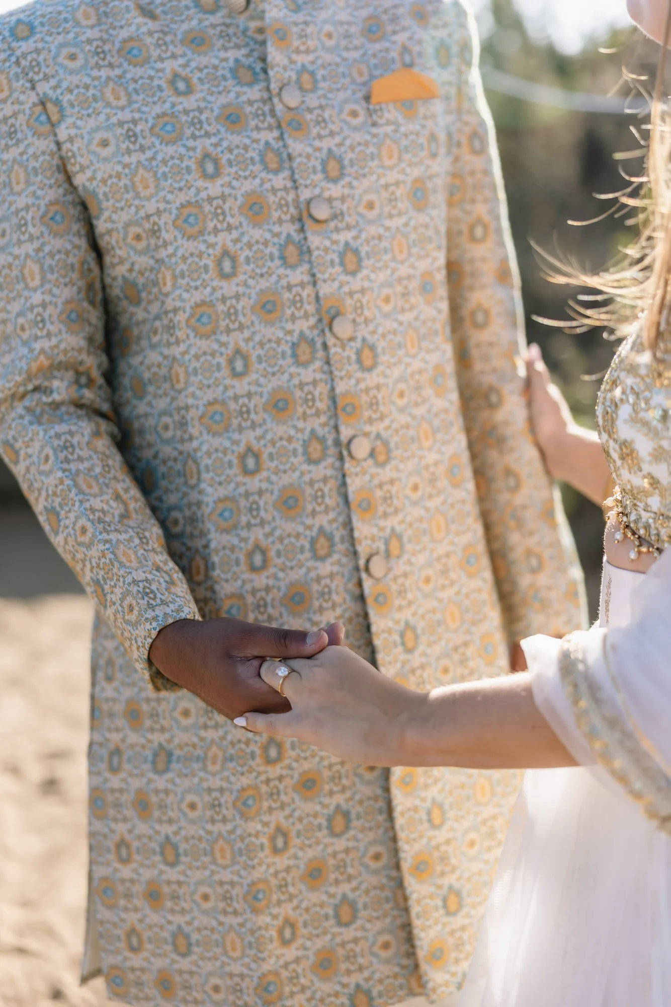 Close-up of a couple holding hands, with the man's hand on top of the woman's, during a wedding ceremony outdoors. The man is wearing a patterned yellow and blue suit, and the woman is wearing a white dress with pearl details.