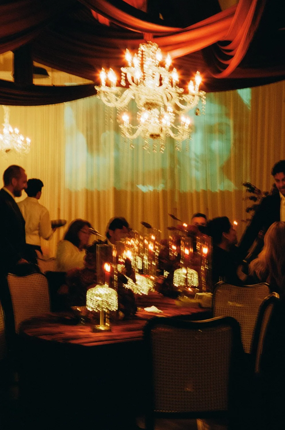 Elegant dinner party with a large chandelier hanging from a decorated ceiling, guests seated at a table with lit star-shaped lights and a projected image of a woman on the wall, creating a warm, festive ambiance.