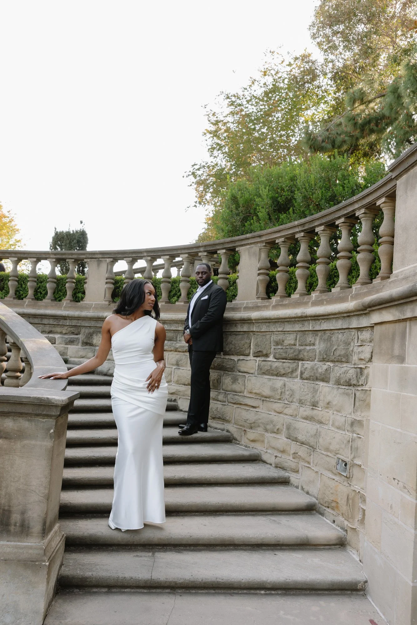 A woman in a white strapless gown leaning on a stone balustrade on a staircase, with a man in a black suit standing behind her on the stairs, all outdoors with trees and sunlight in the background.