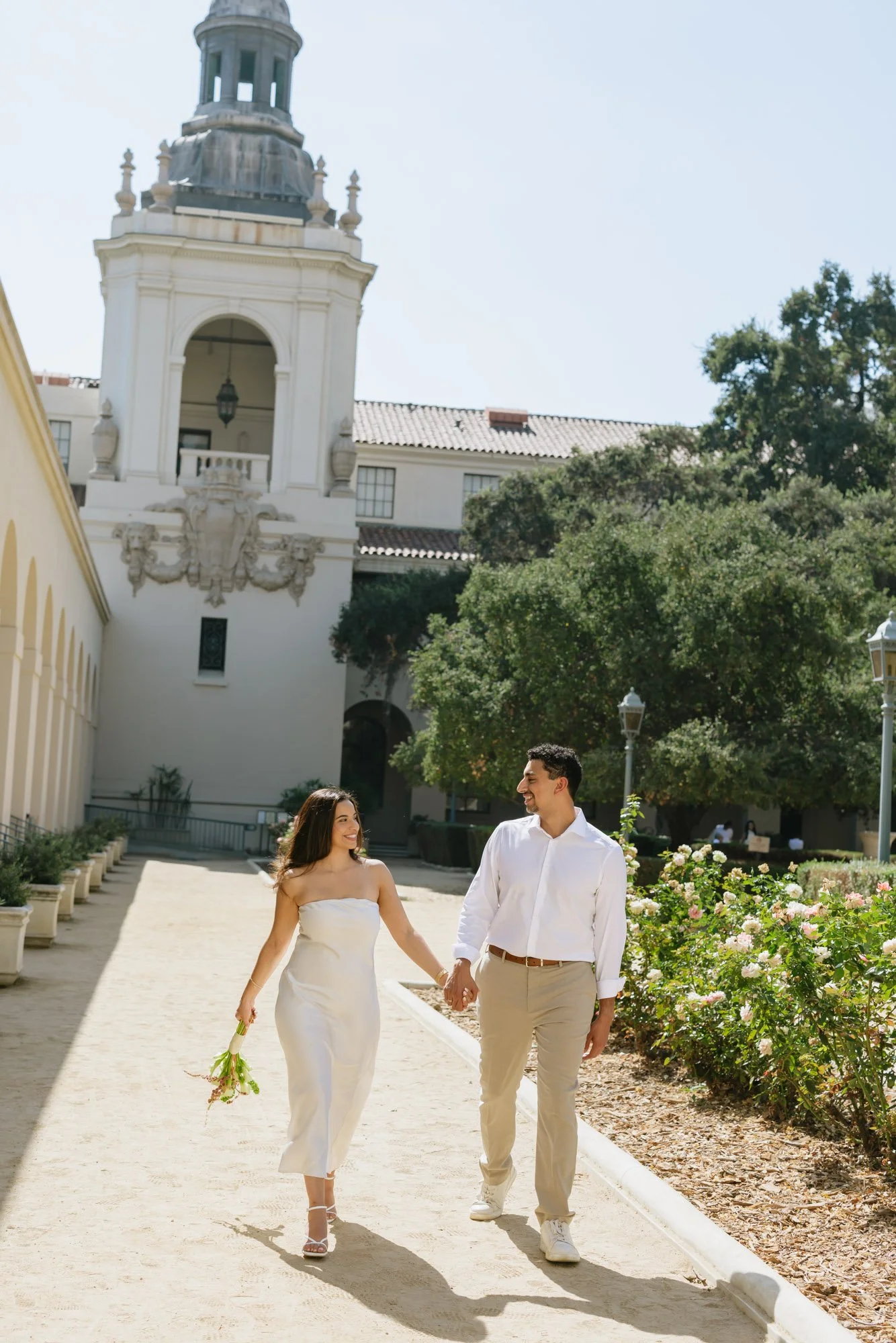 A bride and groom walking hand in hand outdoors on a sunny day, surrounded by flowers and greenery, with a historic building with a clock tower behind them.
