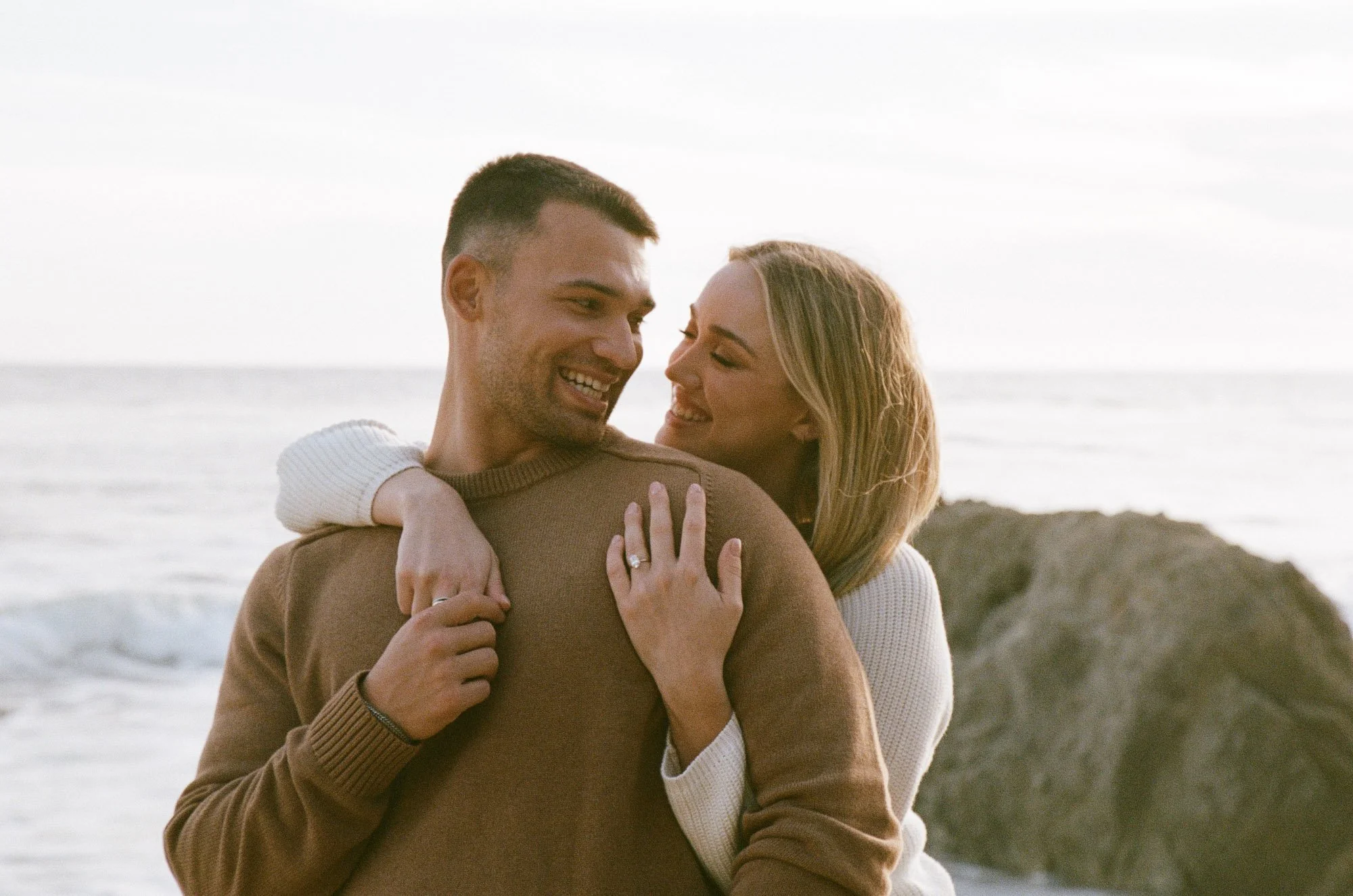 A man and woman embracing on a beach, with the ocean and cliffs in the background, smiling and looking happy.