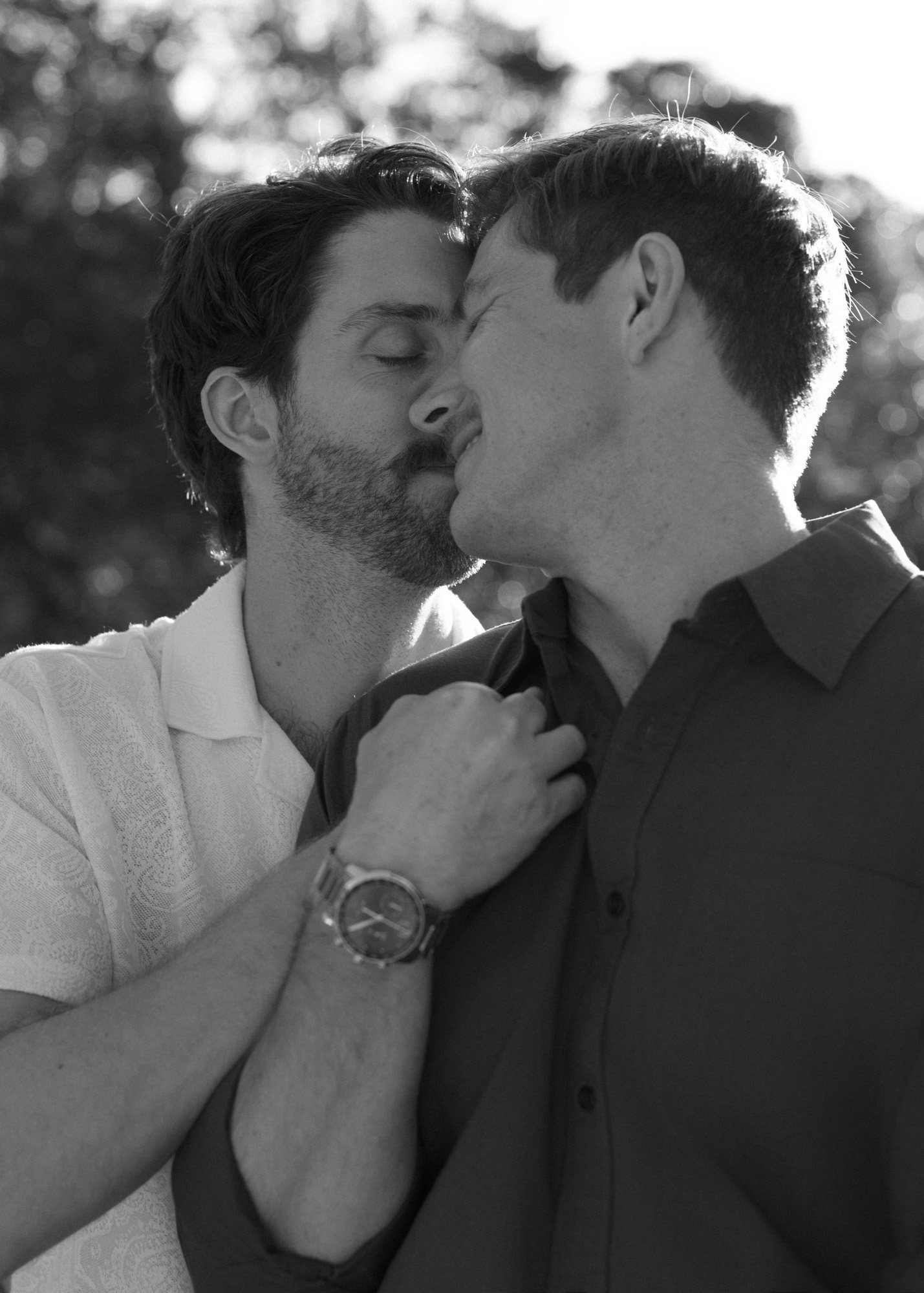 Two men are close together with foreheads touching, sharing an intimate moment outdoors. One has dark hair and a beard, the other has lighter hair. The background is blurred with trees.