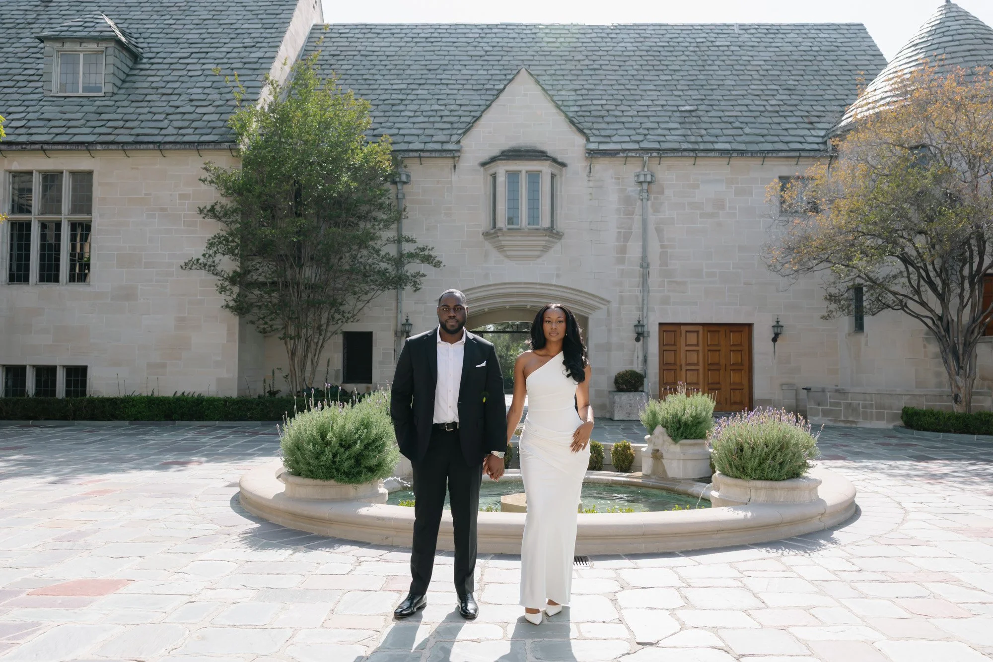 A man and woman dressed in formal attire, standing outside in front of a stone mansion with a fountain and garden, during daytime.