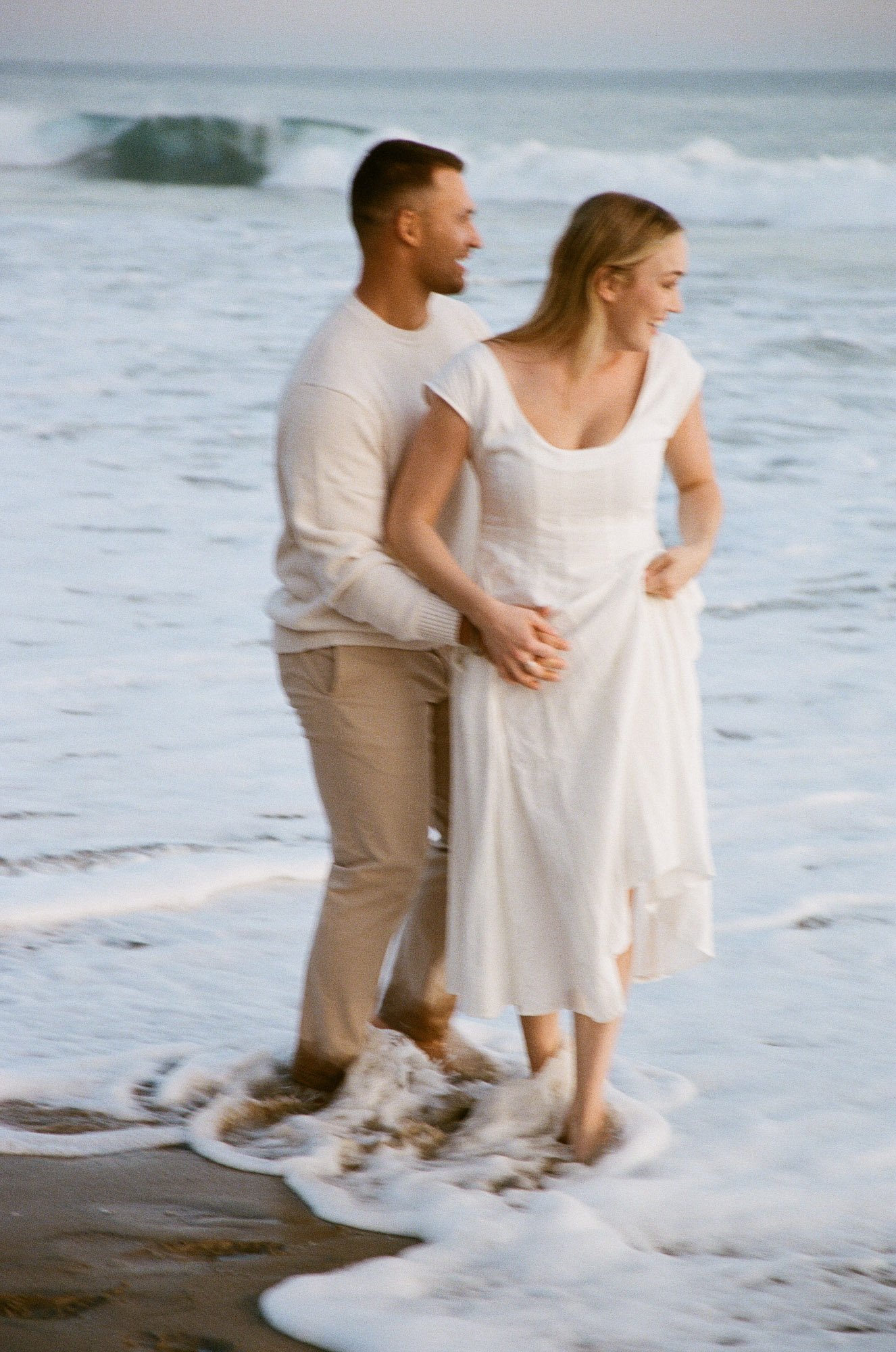 A couple walking hand in hand along the shoreline at the beach, with waves gently crashing around their feet, smiling and enjoying each other's company.