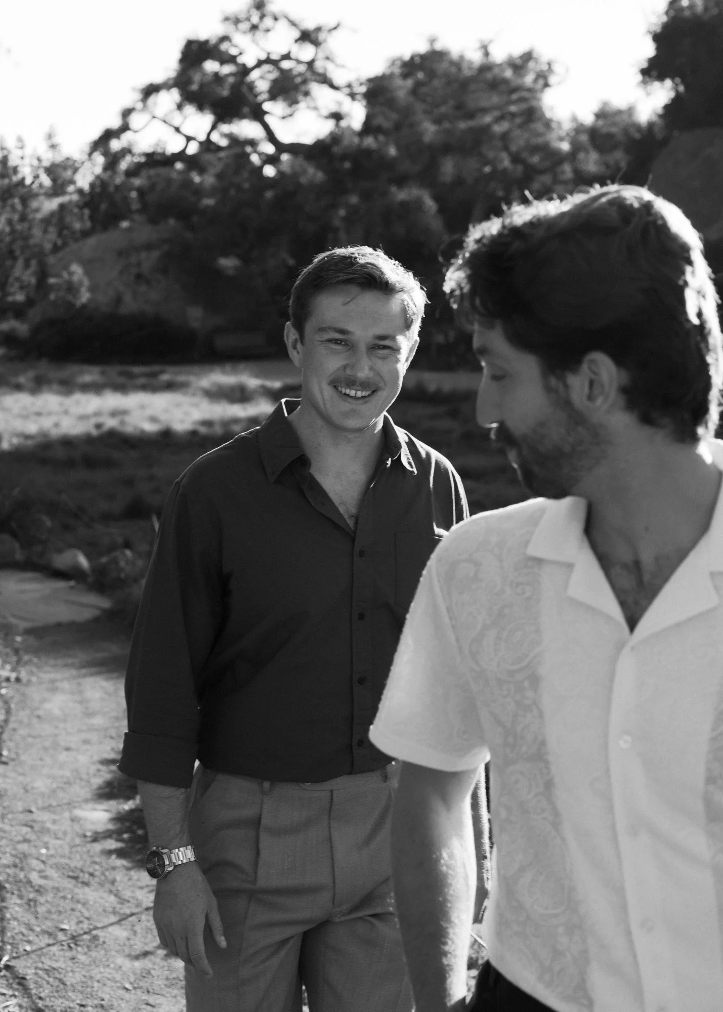 Two men standing outdoors, one smiling and looking at the camera, the other with his head turned down. Trees and a grassy hill are in the background, captured in black and white.