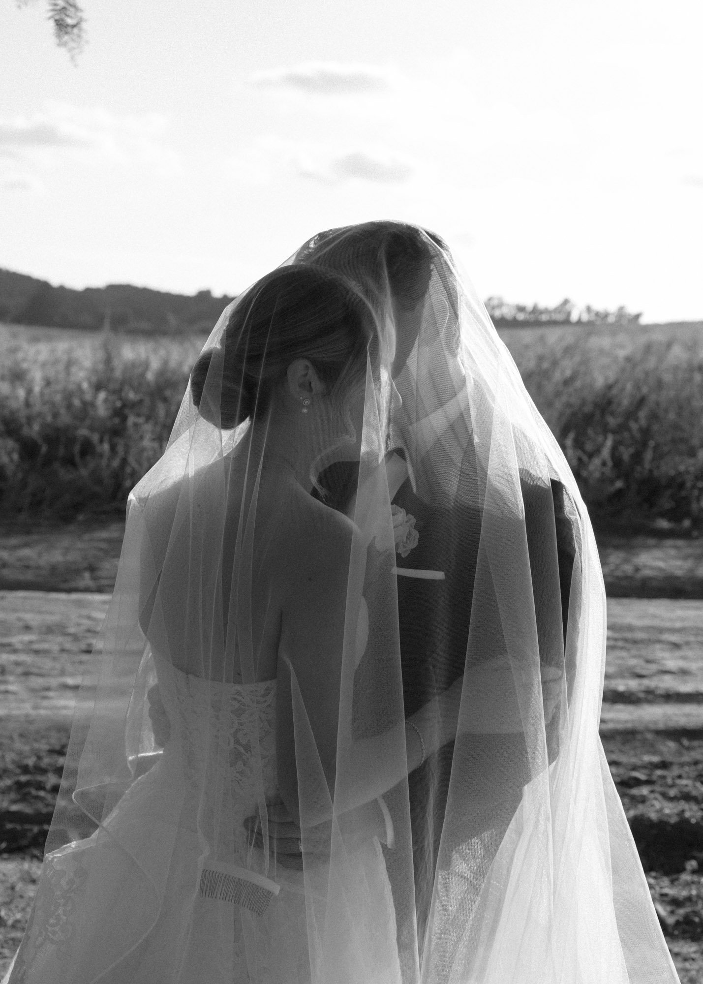 A black-and-white photo of a bride and groom embracing outdoors, with the bride wearing a veil and lace dress, and the groom in a suit, in a field with a hill and cloudy sky in the background.