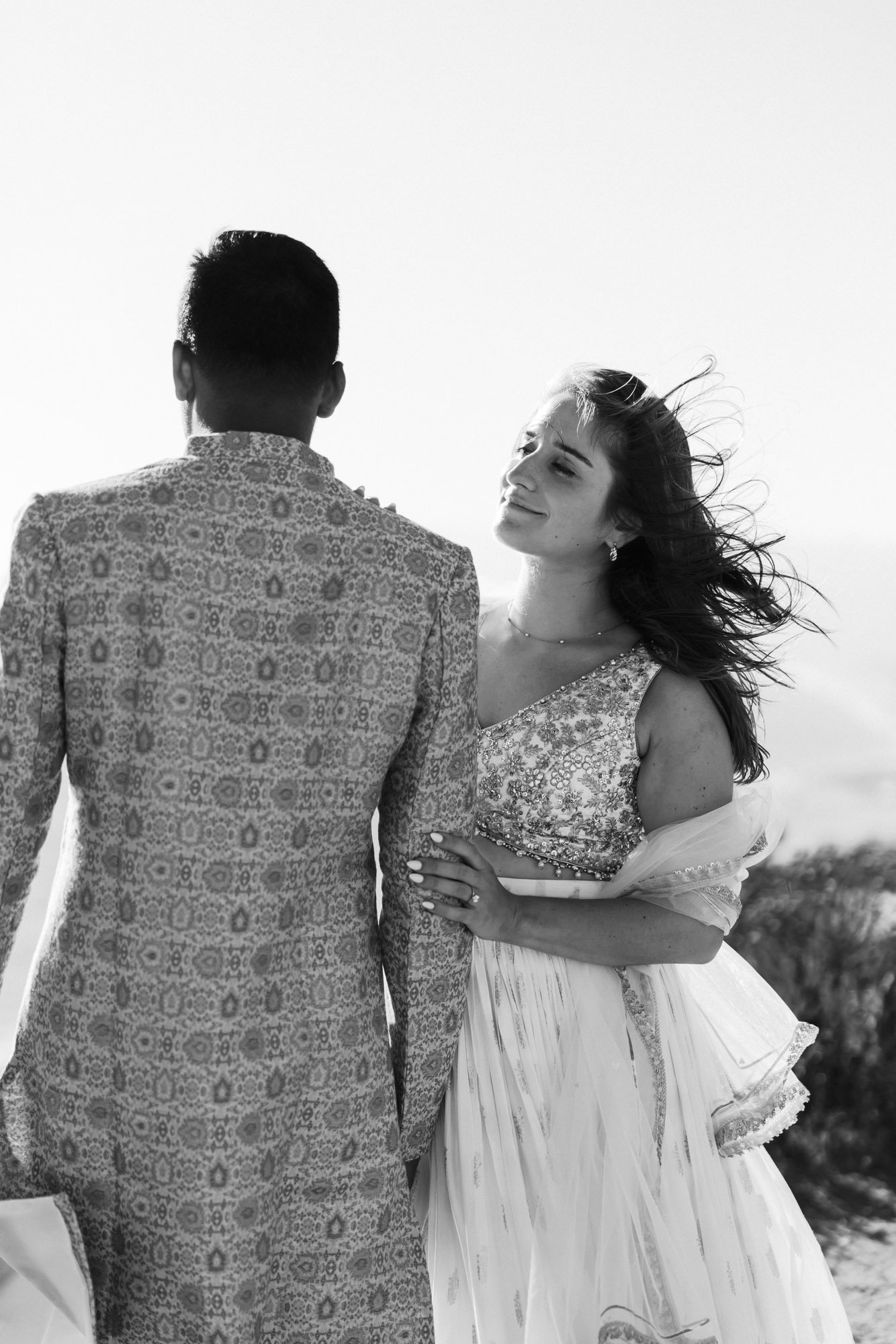 A black and white photo of a woman with long, flowing hair looking tenderly at a man whose back is to the camera. The man wears a patterned jacket, and the woman wears a dress with intricate embroidery and a full skirt, suggesting a formal or special