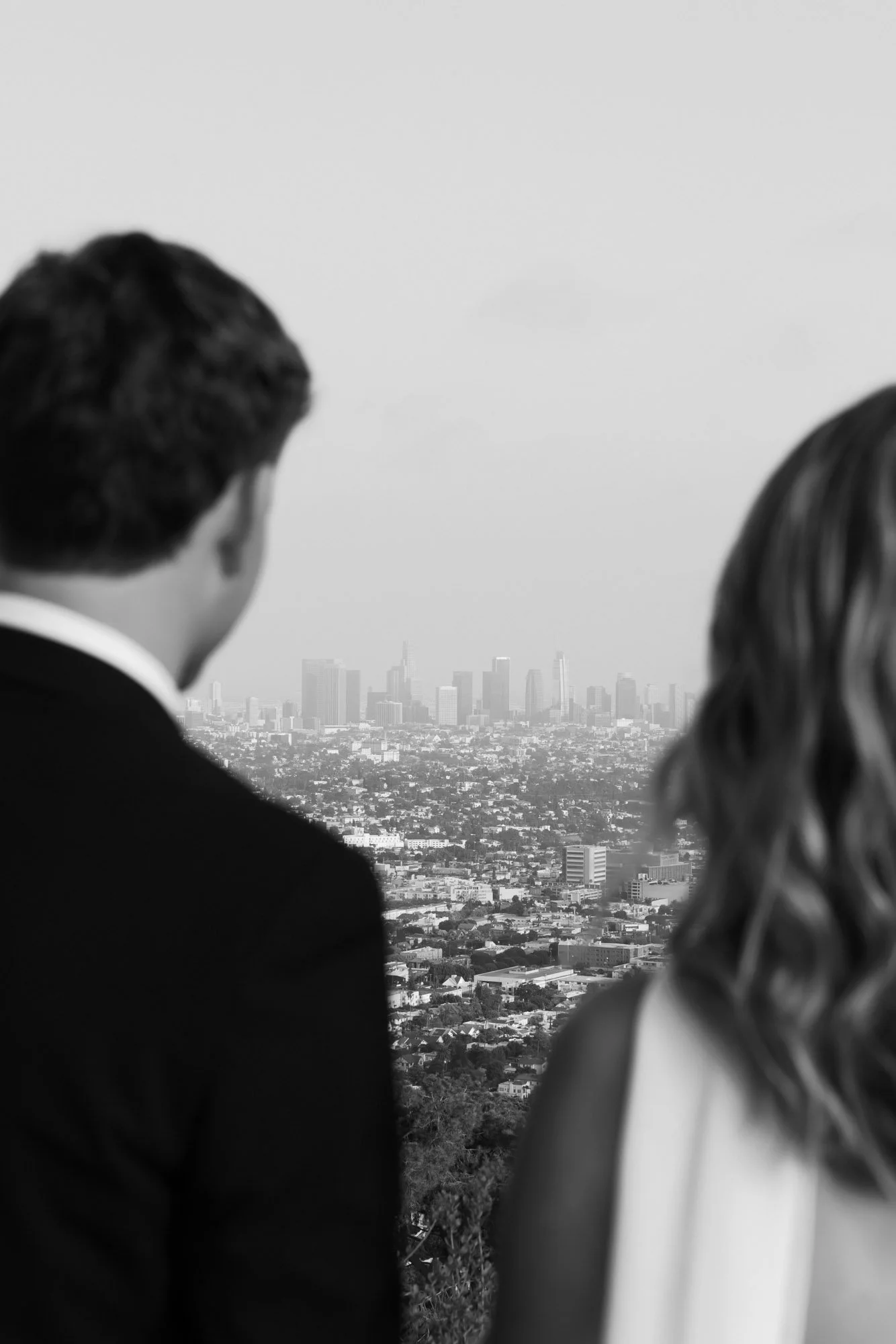 Black-and-white photo of two people, a man and a woman, seen from behind, looking out over a city skyline from a high vantage point.