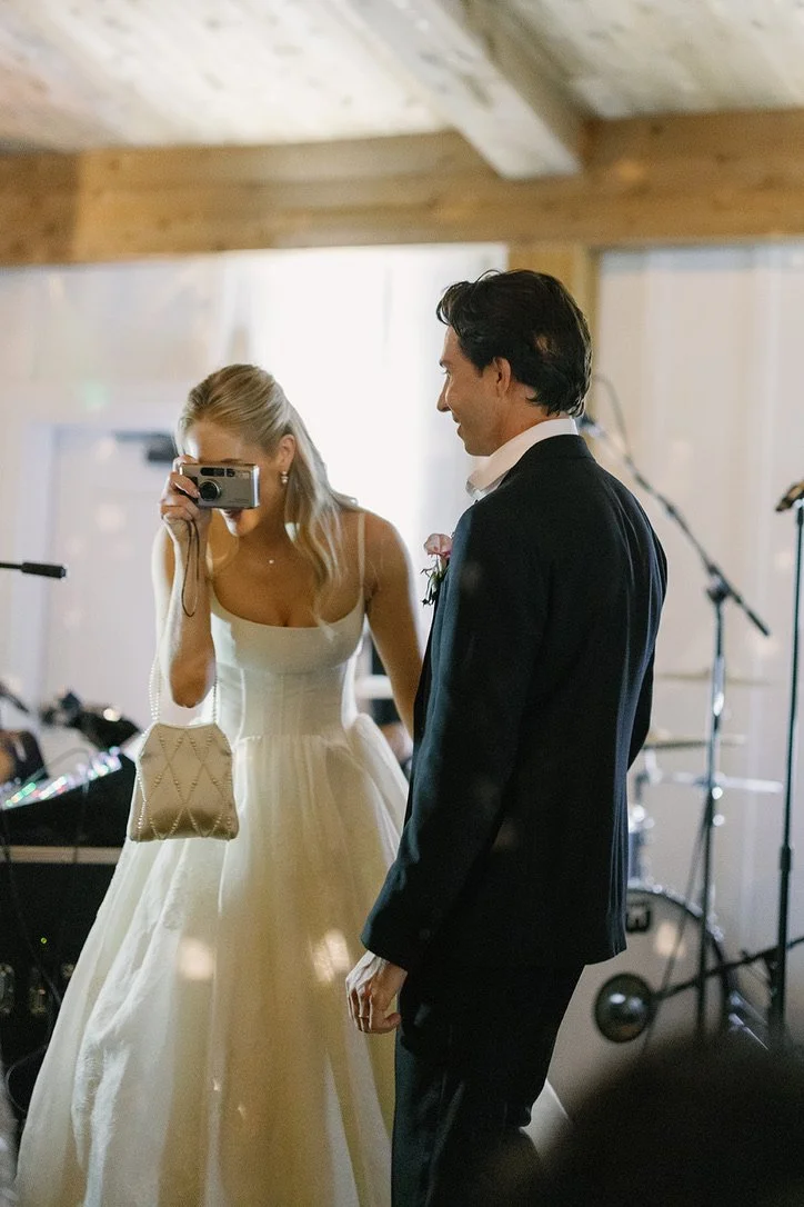 A bride and groom at a wedding reception, with the bride taking a photo of the groom. The bride is in a white wedding dress and the groom is in a dark suit.