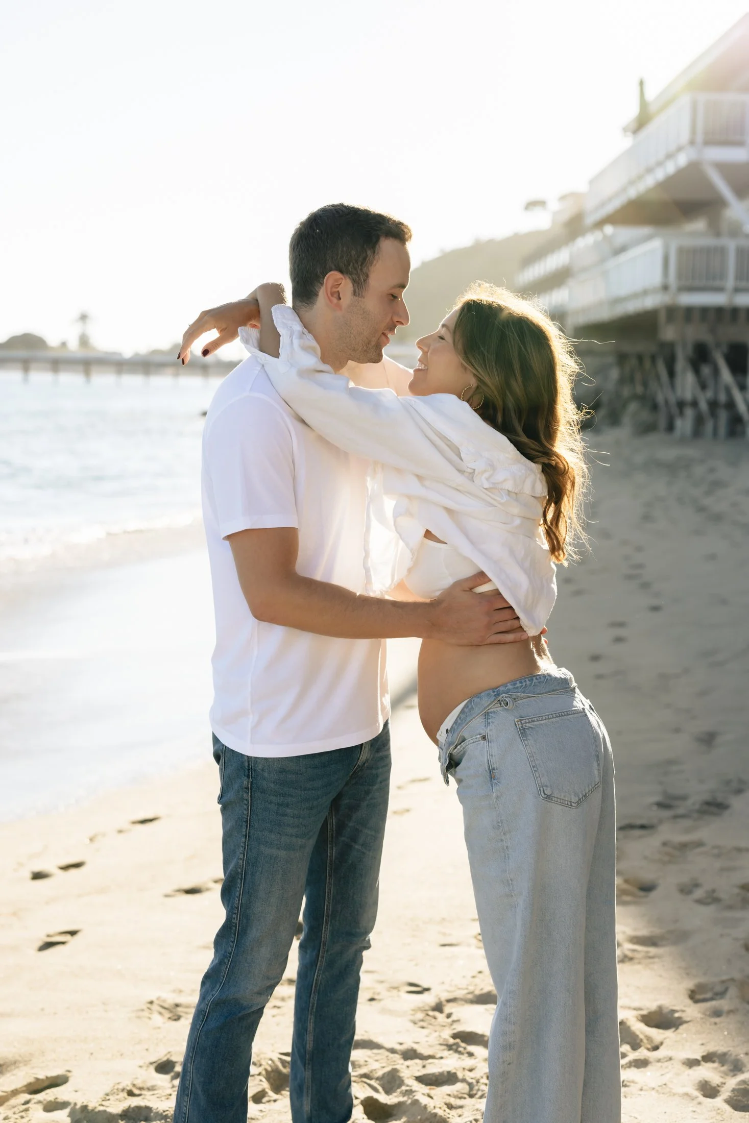 A young couple is embracing on the beach during sunset, with the man holding the woman and smiling at her. The woman is smiling back, with her arms around his neck. In the background, there are houses on stilts and a pier extending into the water.