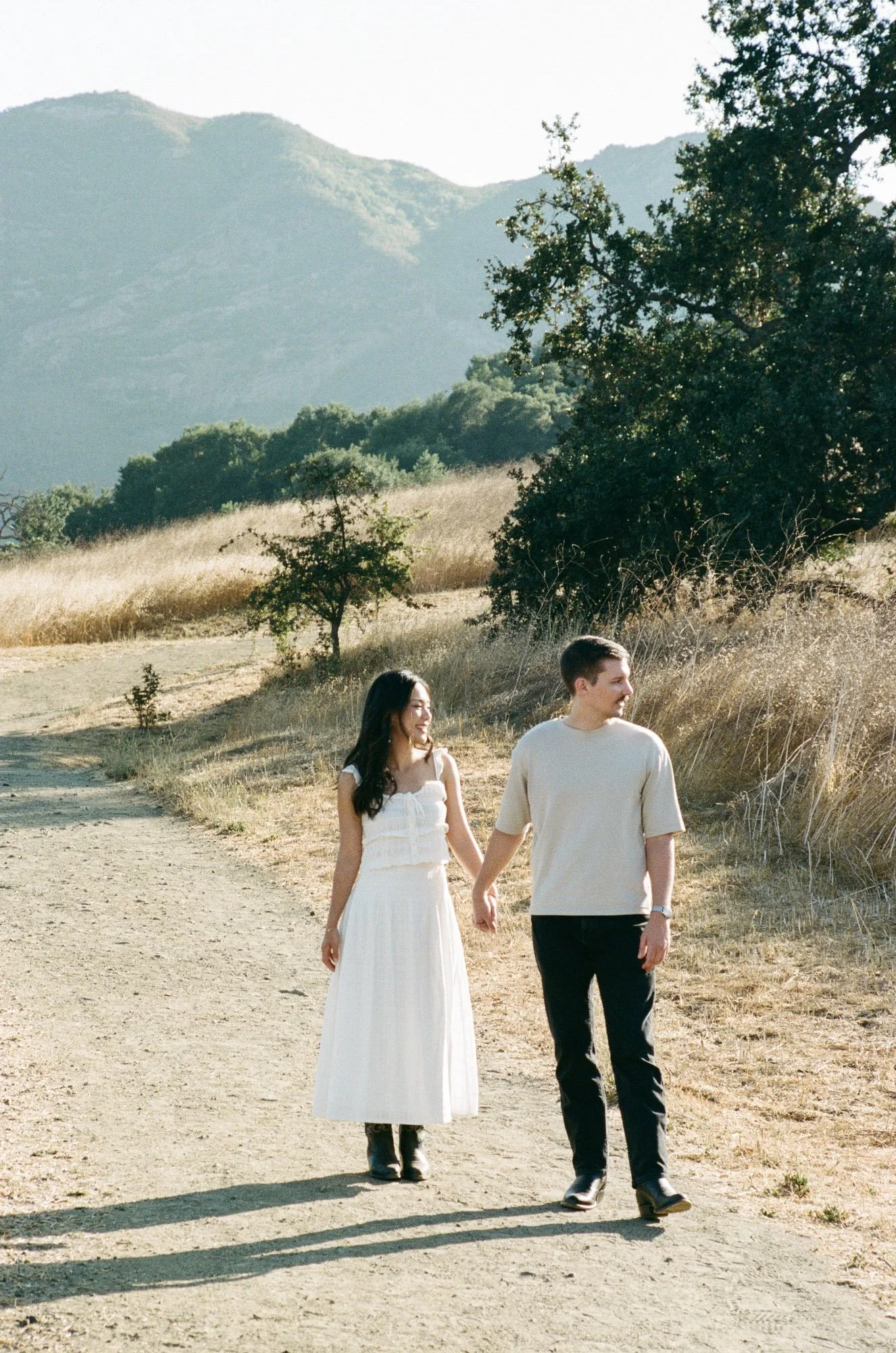 A young couple walking hand-in-hand on a dirt path in a natural outdoor setting with mountains and trees in the background. The woman is wearing a white dress and black boots, and the man is dressed casually in a beige shirt and dark pants.