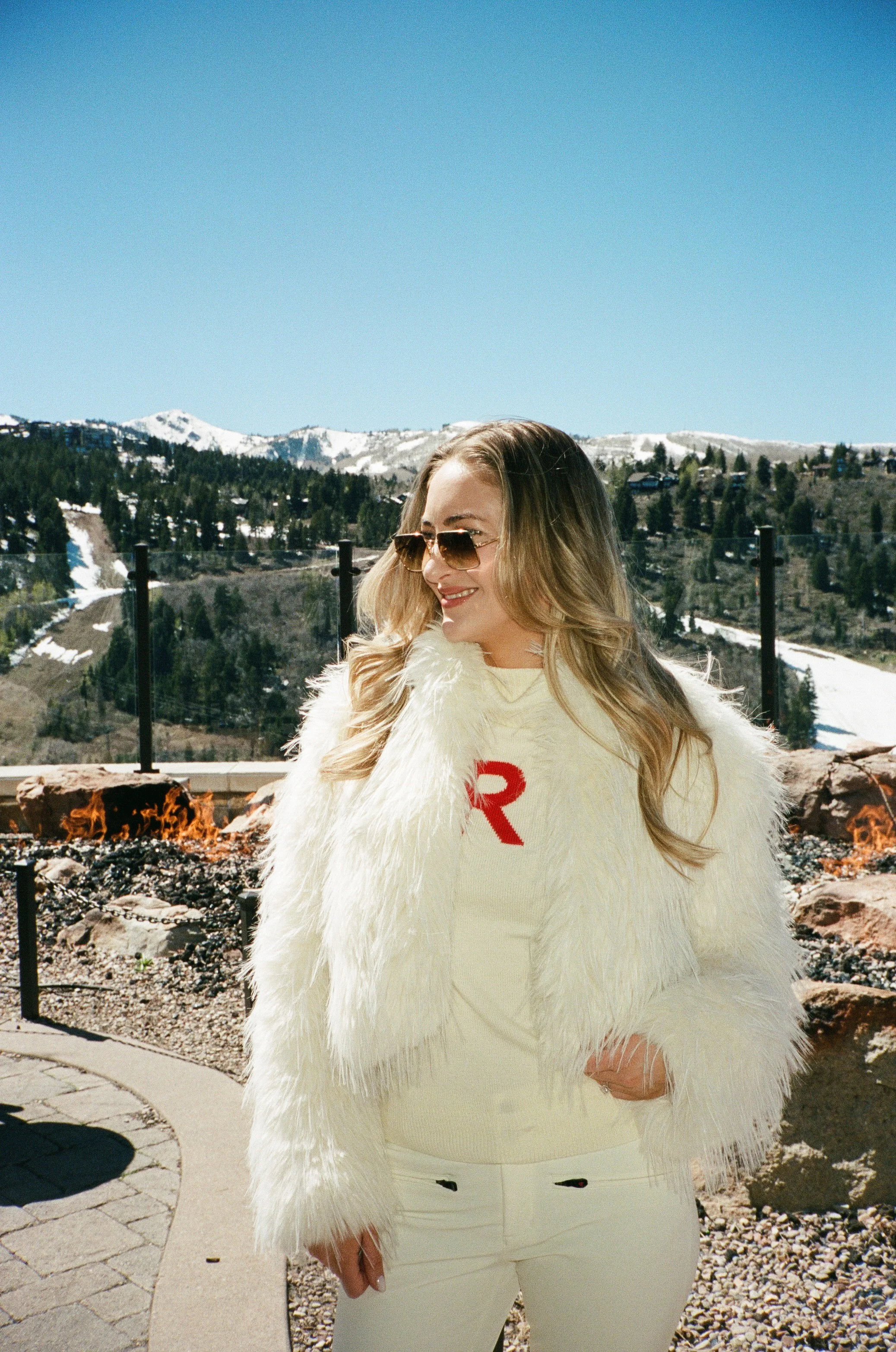 A woman with blonde hair wearing sunglasses and a cream-colored fur coat over a matching outfit stands outdoors with a mountain landscape in the background. There is a fire pit with flames in front of her.