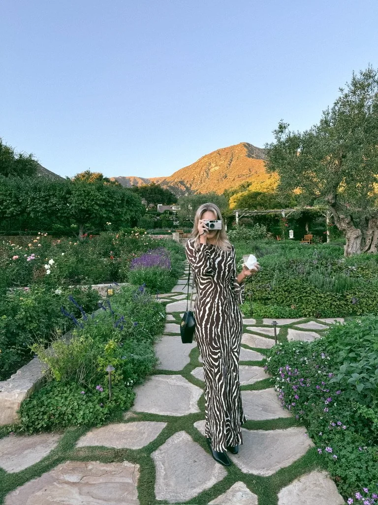 A woman in a zebra-print dress taking a photo with a camera on a stone pathway in a lush garden with colorful flowers, trees, and mountains in the background.