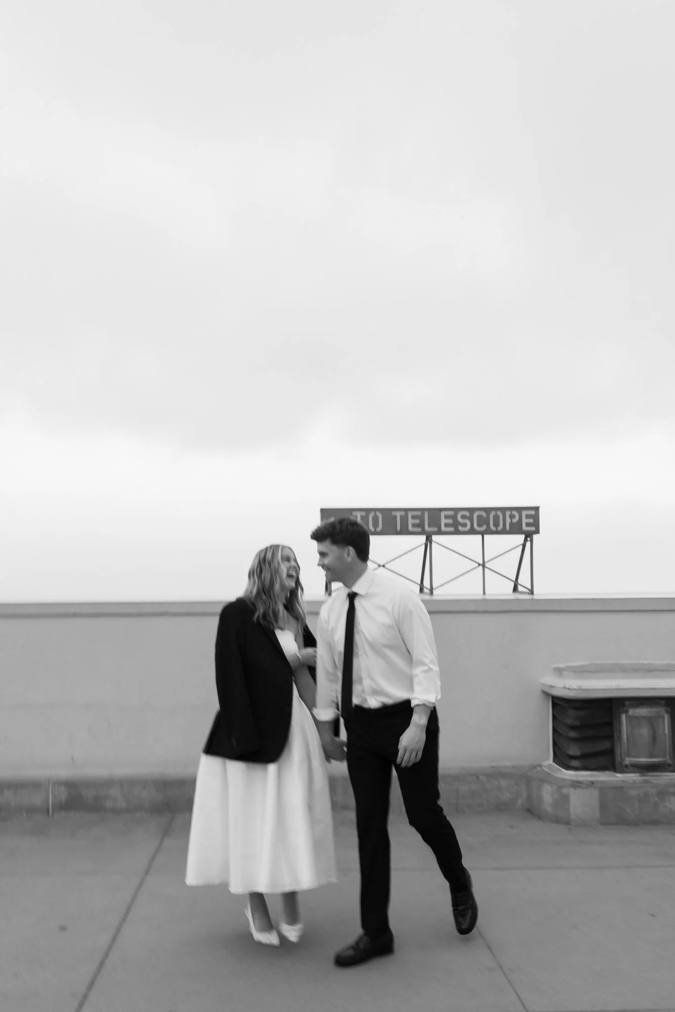 A black and white photo of a man and woman standing together outdoors, smiling and holding hands. The woman is wearing a long dress and blazer, and the man is in a shirt and tie. There is a sign in the background that reads "TO TELESCOPE."