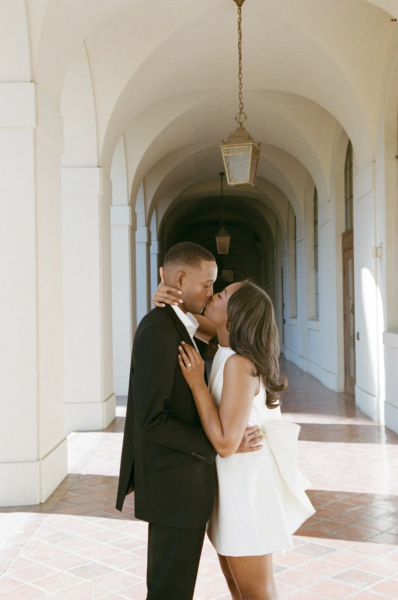 A couple sharing a kiss in an arched outdoor corridor, with lanterns hanging from the ceiling.