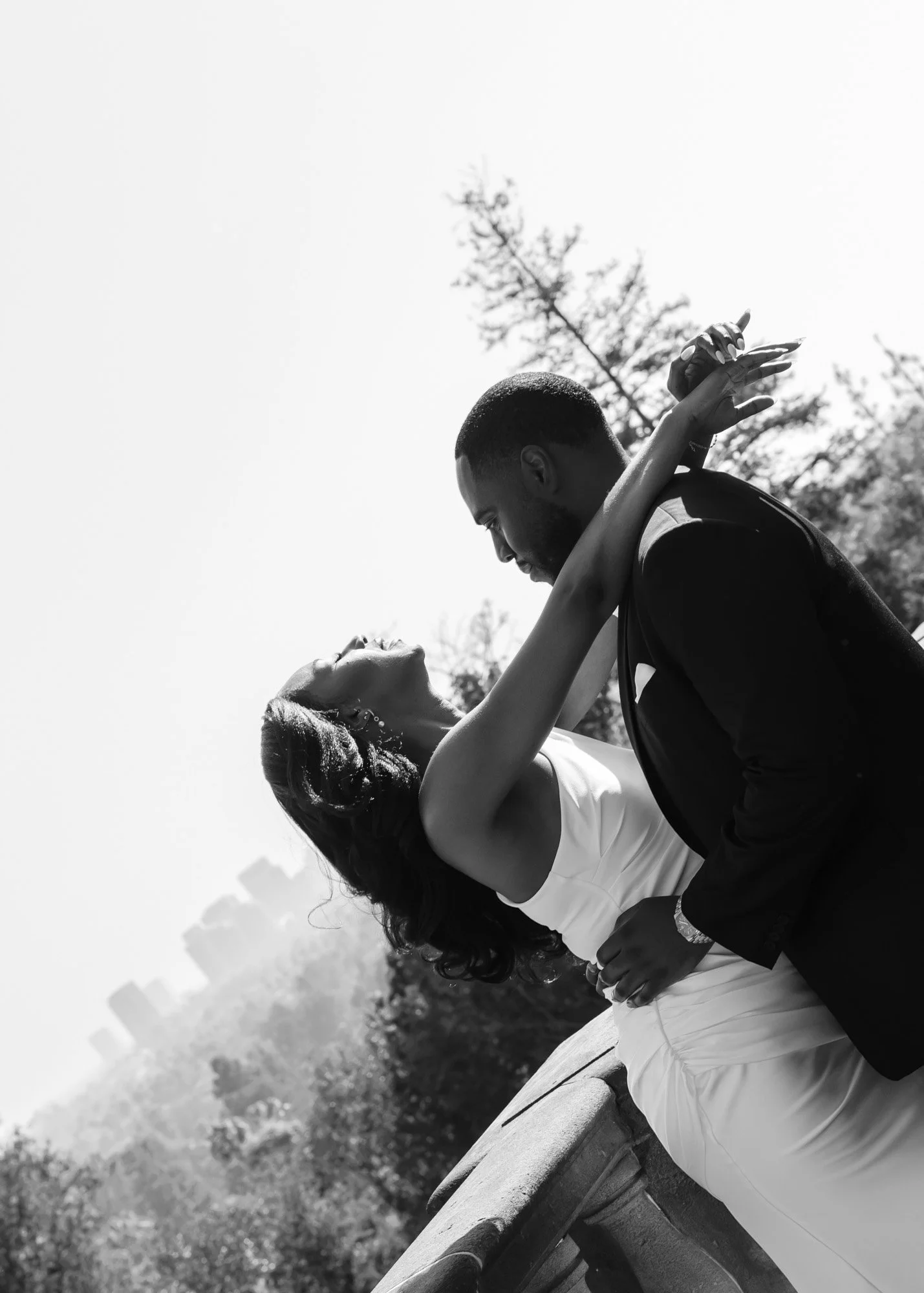 A couple dances outdoors on a sunny day, with the man holding the woman as she leans backward, eyes closed, smiling. The background includes trees and a distant city skyline.