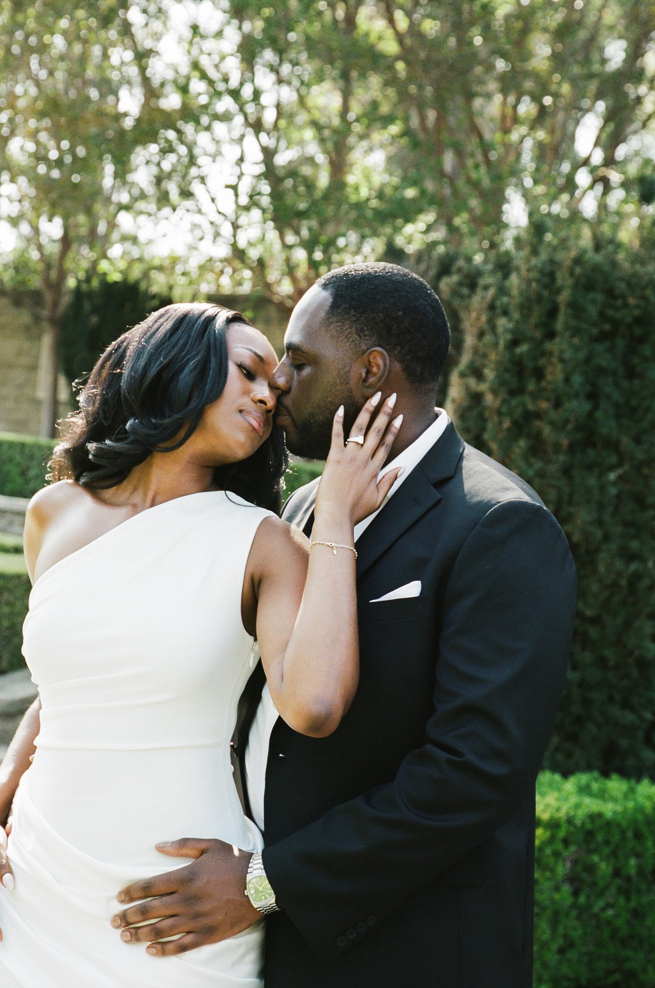 A couple dressed in formal attire, with the woman in a white dress and the man in a black suit, sharing an intimate moment outdoors with a background of trees and greenery.