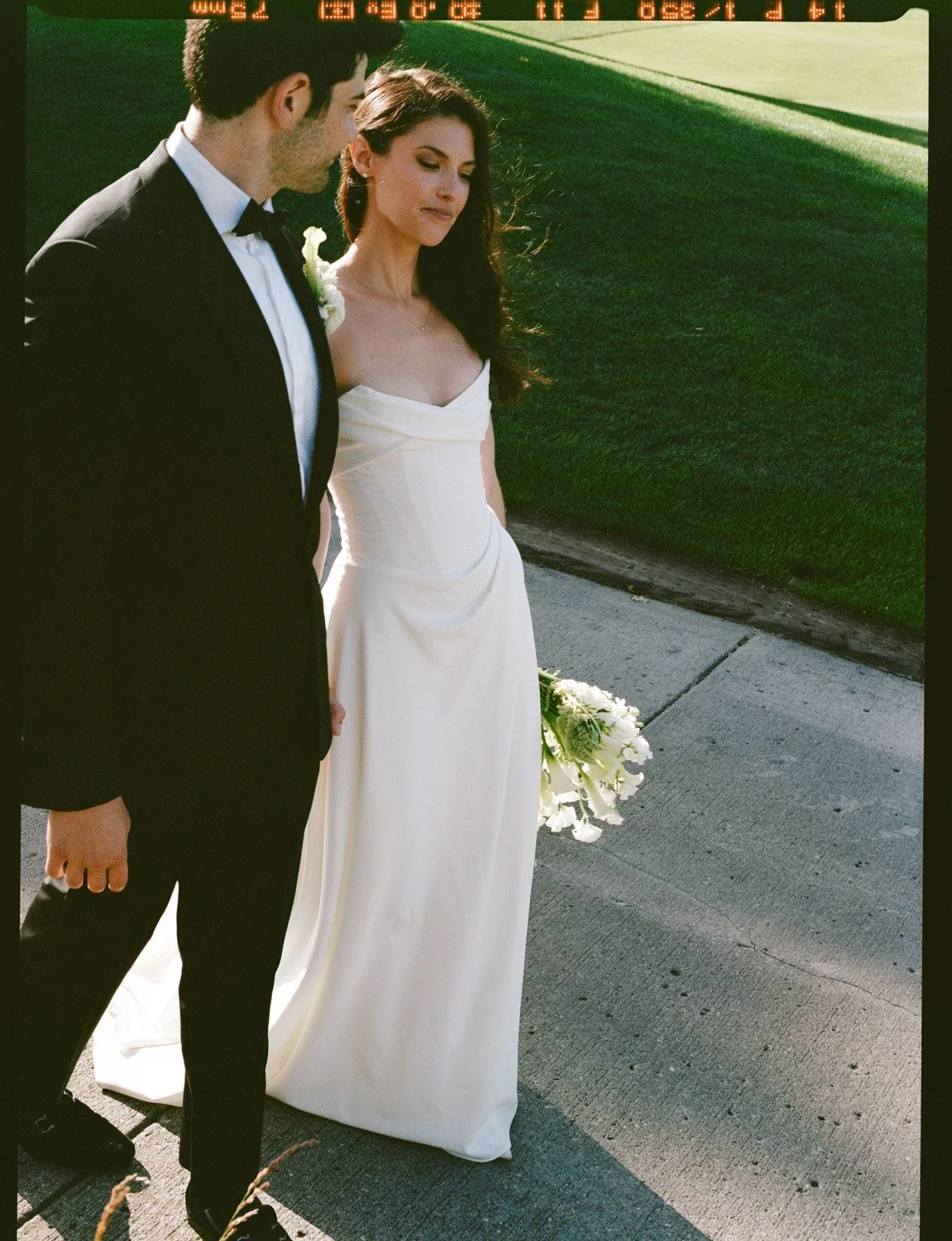 A bride and groom standing outdoors on a sidewalk during their wedding. The groom is wearing a black tuxedo and the bride is in a white wedding gown holding a bouquet of white flowers. The sun is low, casting warm light.