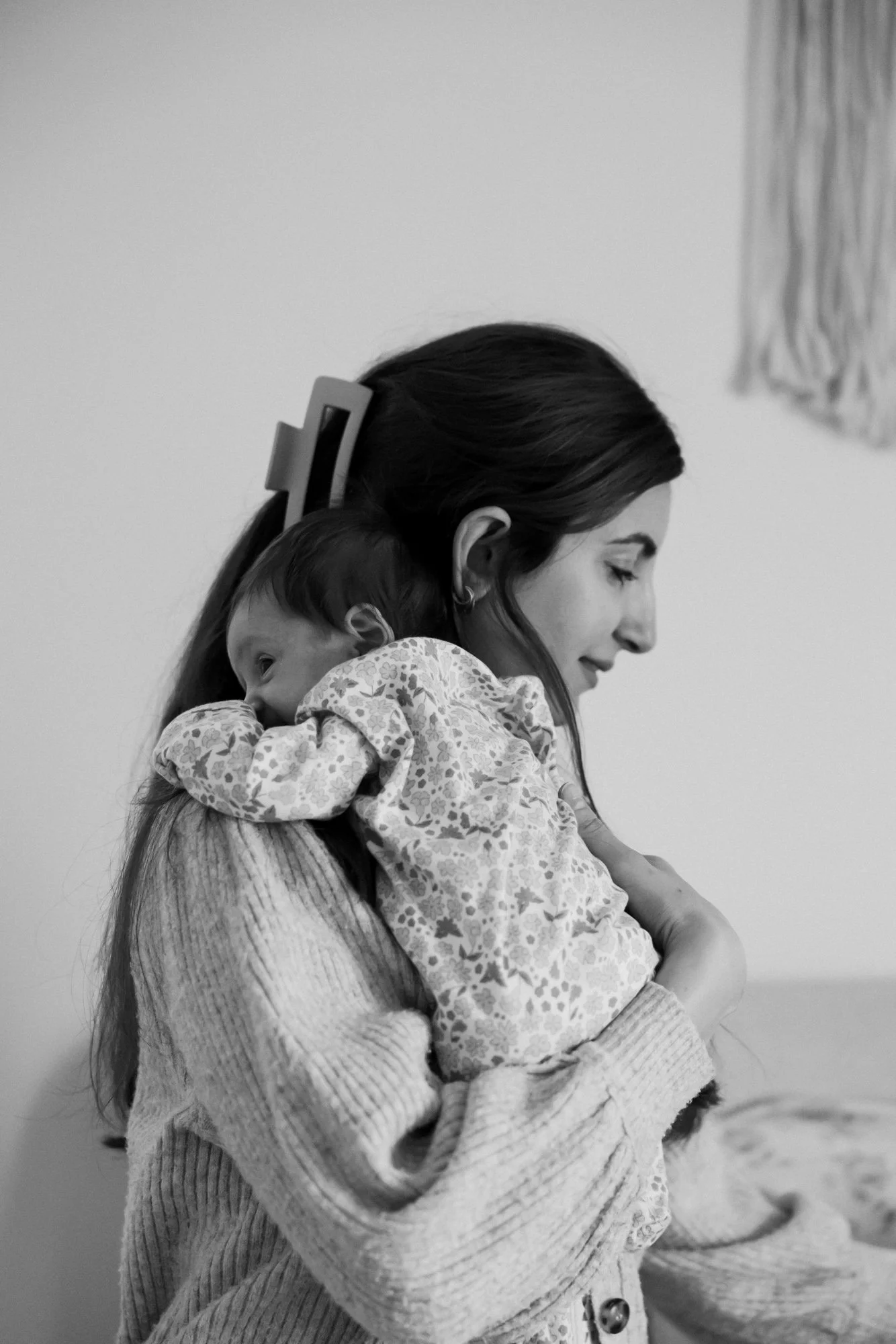 A woman with long hair, hair clip, and hoop earrings, holding a young girl with dark hair and a floral-patterned dress, both with eyes closed, in a close embrace. Black and white photograph.
