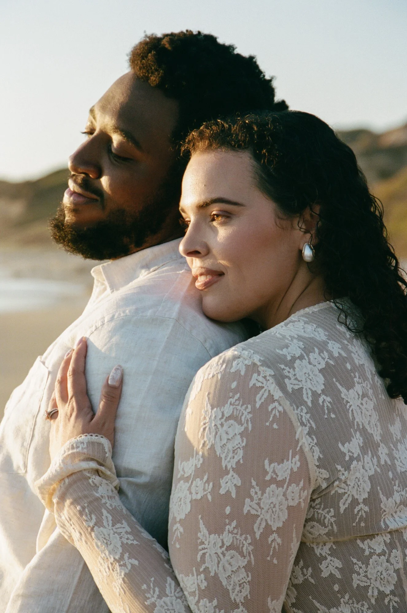 A couple embracing on a beach at sunset, with the woman leaning on the man’s shoulder and the man's eyes closed, both smiling gently.