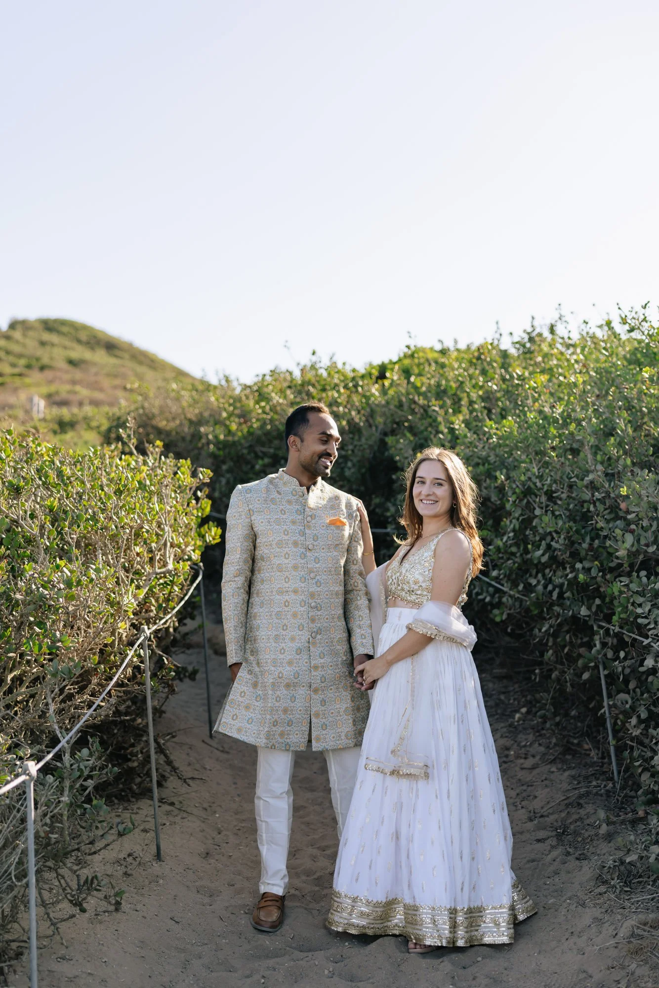 A couple in traditional Indian attire stands on a dirt path surrounded by green bushes, holding hands and smiling at each other with a clear sky and a hillside in the background.