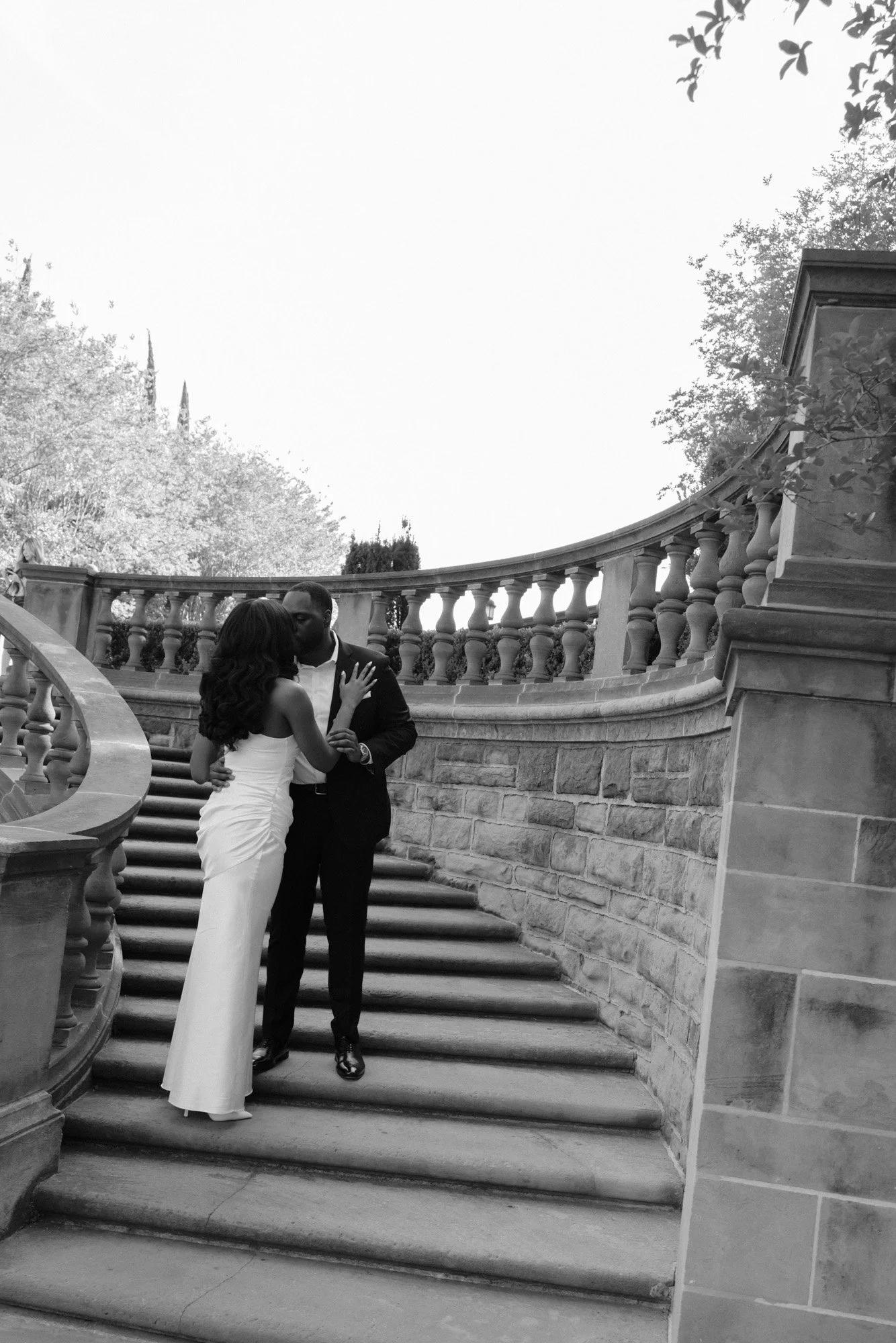 A couple dressed in wedding attire sharing a kiss on a staircase with stone balustrades outside, surrounded by trees.
