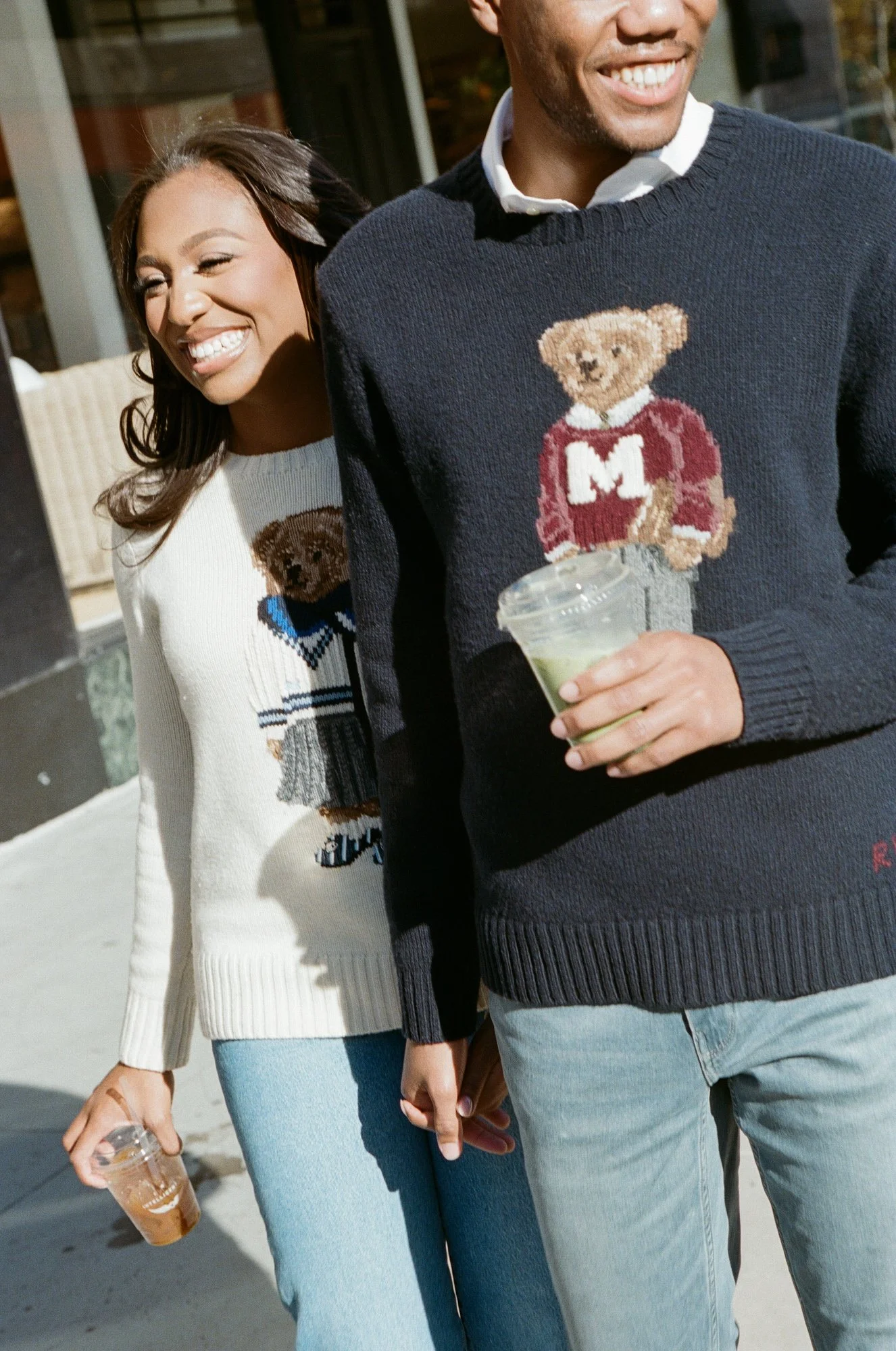 A man and woman holding hands, wearing matching sweaters with teddy bear designs and holding drinks, walking outdoors in daylight.