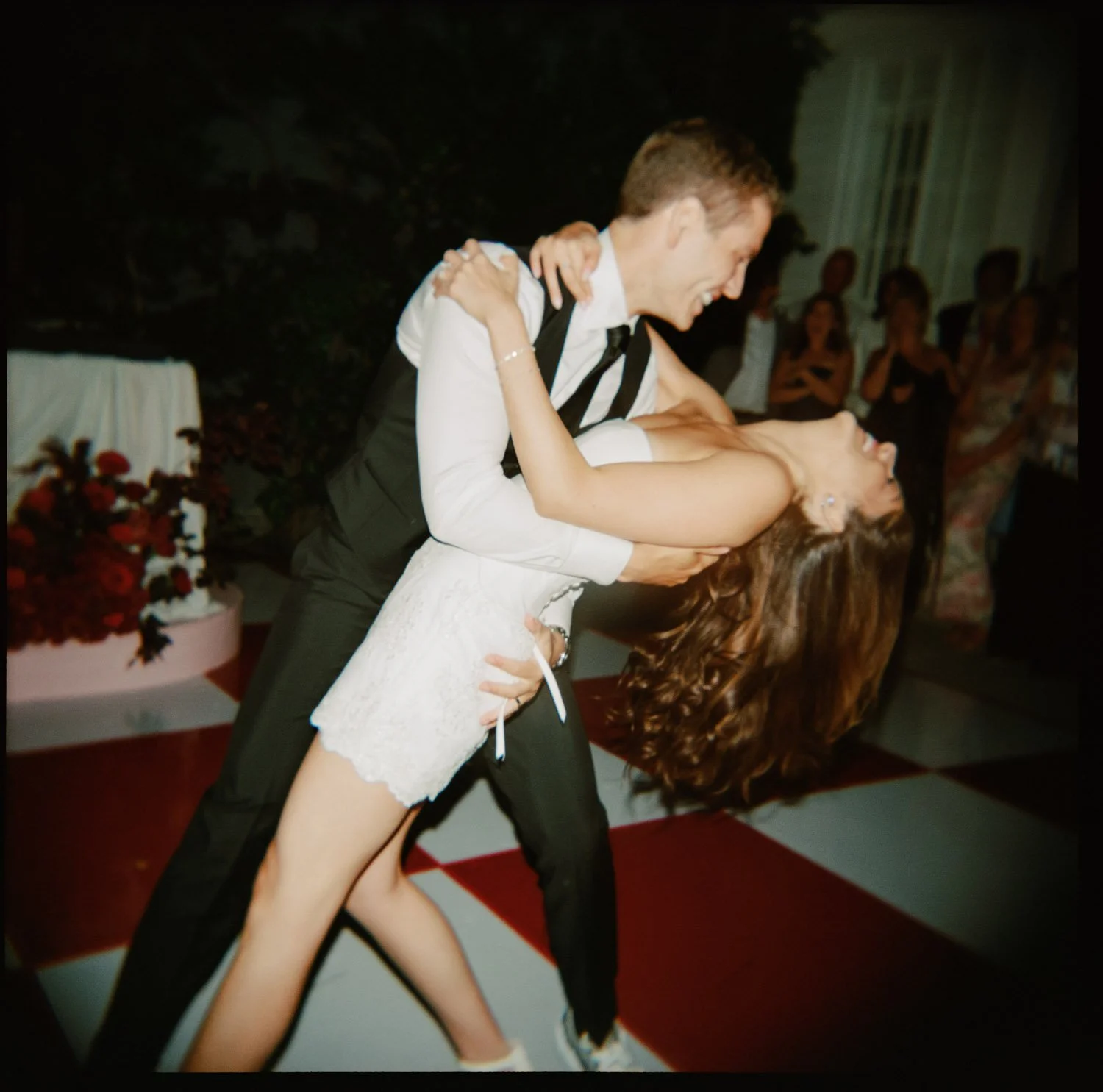 A couple dancing closely at a wedding reception, with guests watching in the background and a decorated table with flowers nearby.
