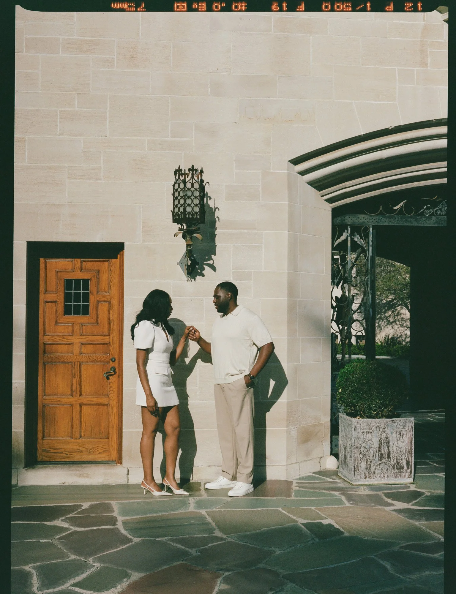 A man and woman standing outside a building, holding hands and engaging in conversation, with a beige stone wall, wooden door, and decorative ironwork in the background.