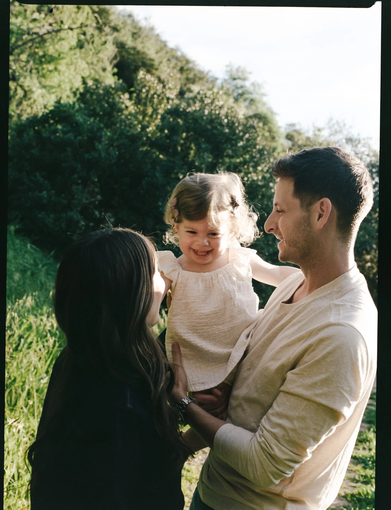 A man is holding a smiling young girl outdoors in a green park with trees and grass, during daytime.
