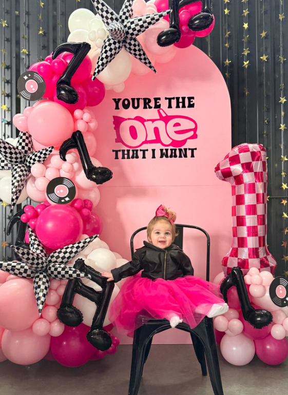A young girl sitting in a black chair, wearing a black jacket and a pink tutu, in front of a pink backdrop with the text "You're the one that I want." The background is decorated with pink, white, and black balloons, checkered star balloons, record-shaped decorations, and a large pink and white checkered number one balloon.